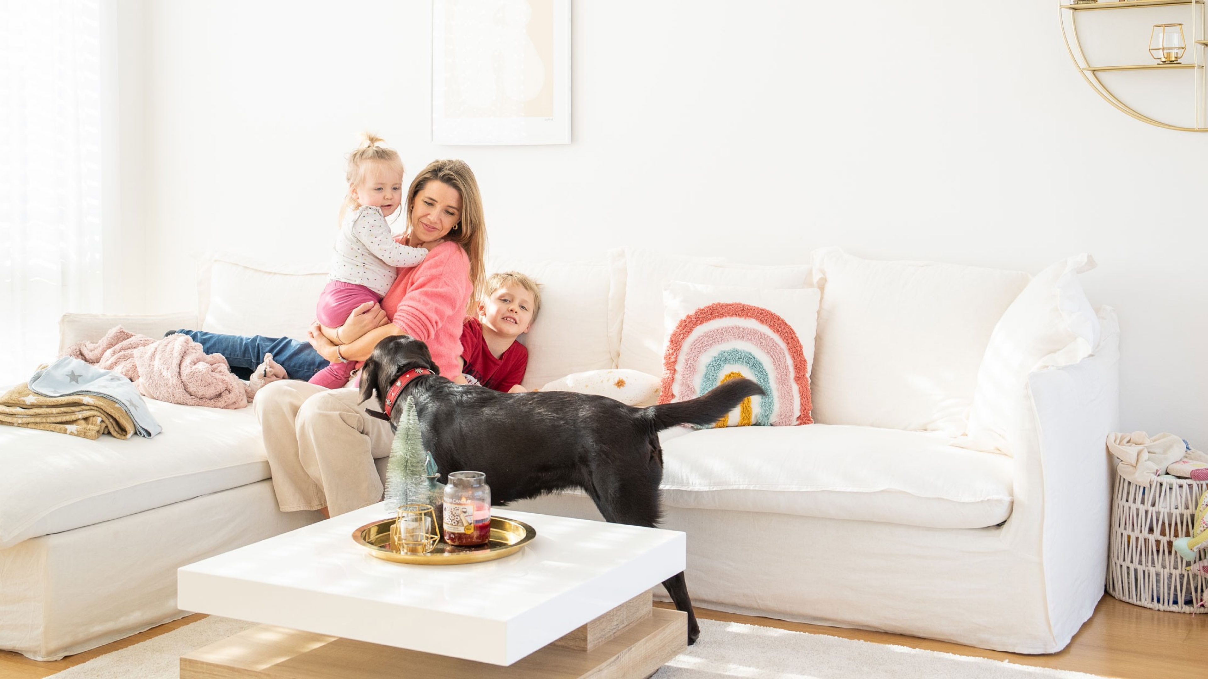 A woman sits on the sofa with her two children. A Labrador stands in front of them.