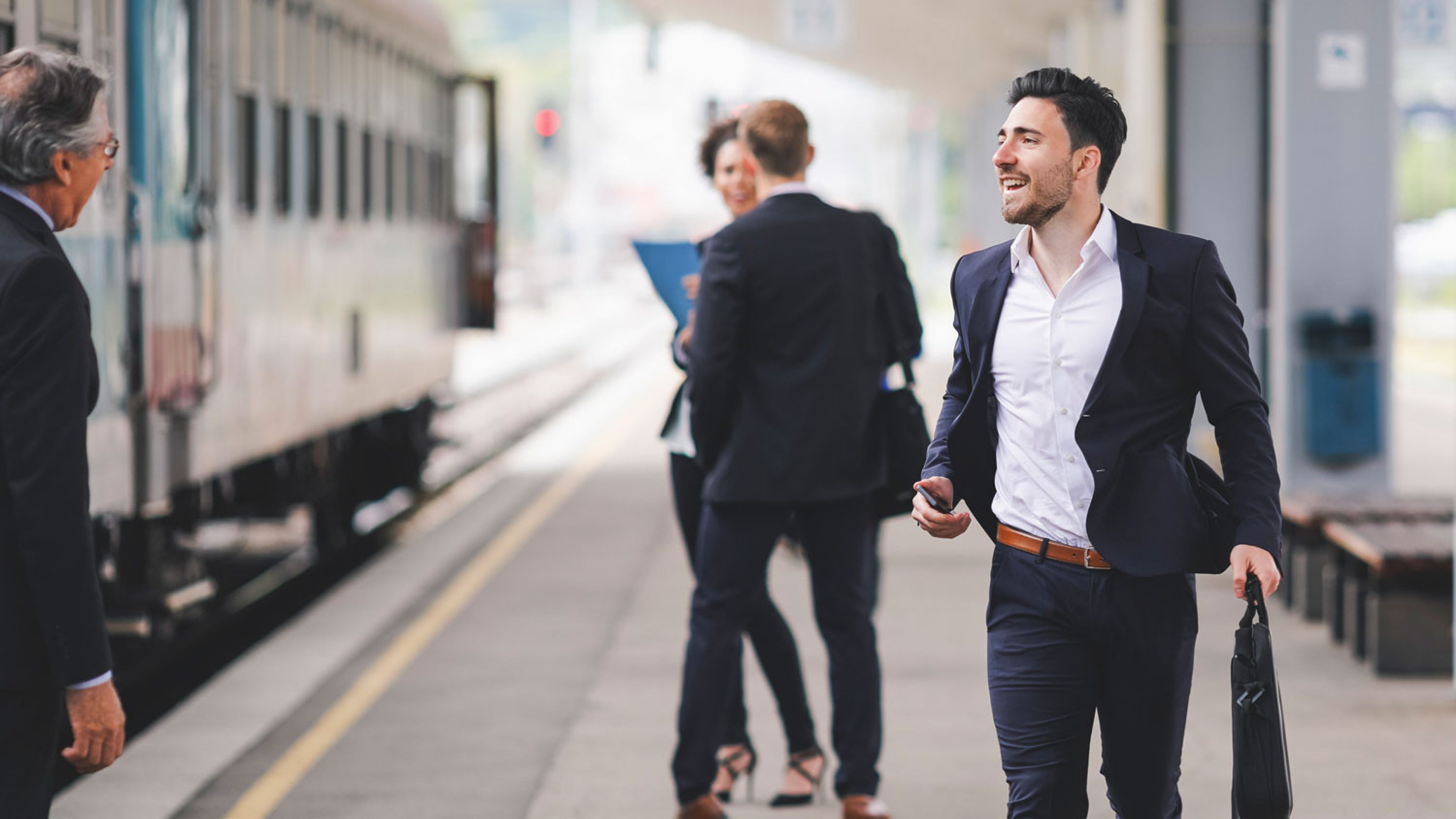 Some people in business clothes are on a platform. A smiling man wearing a suit and holding a briefcase approaches the camera. In the background there are two people talking and a train is pulling in. To the left of the picture is an older gentleman looking at the group of people. He has grey hair and is wearing glasses.