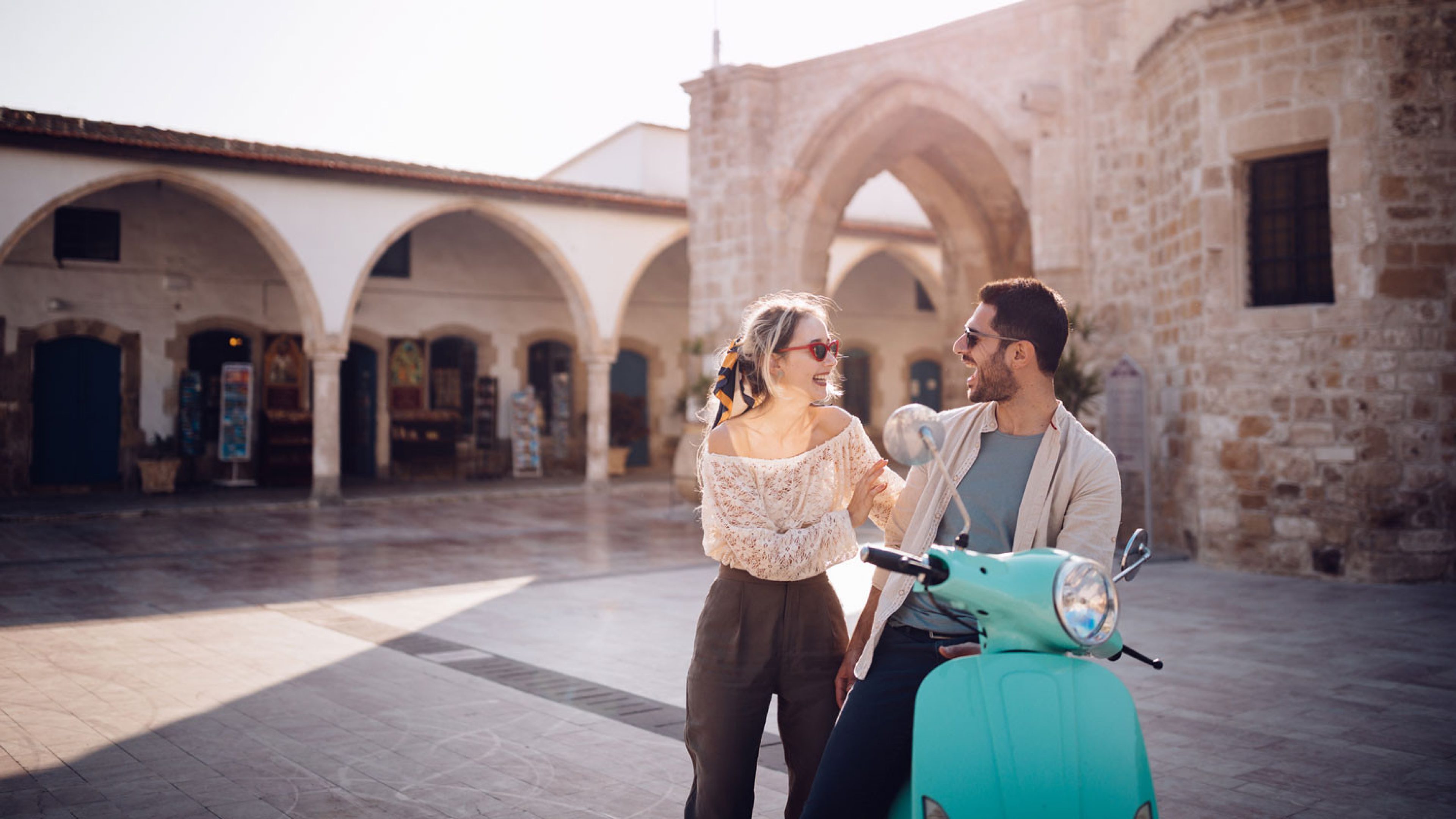 Two people stand next to a turquoise Vespa in an open courtyard with arcades and historic stone buildings in the background. The person on the left is a woman. She wears an off-the-shoulder light top and dark trousers. She is wearing sunglasses and a scarf tied in her blonde hair. The person on the right is a man. He is wearing a light jacket over a blue-grey shirt. He is also wearing sunglasses. Both are laughing. Sunlight in the background casts shadows on the ground.