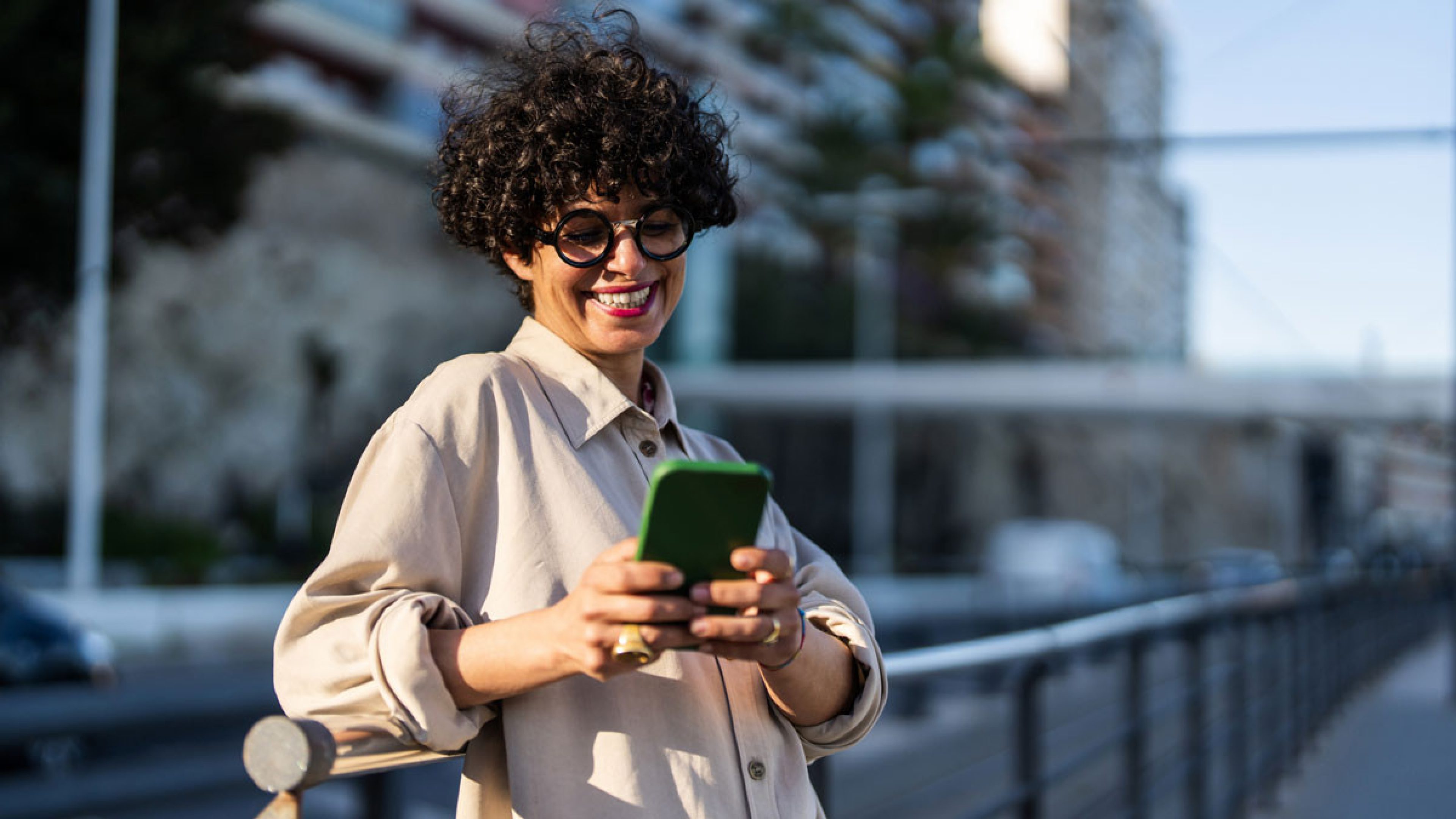 A person with short, curly hair and striking glasses stands outdoors at a metal railing in a modern urban environment. She holds a smartphone in both hands and looks at it as if reading or typing a message. The scene is taken in daylight, with blurred residential buildings, transport infrastructure and glass facades in the background.