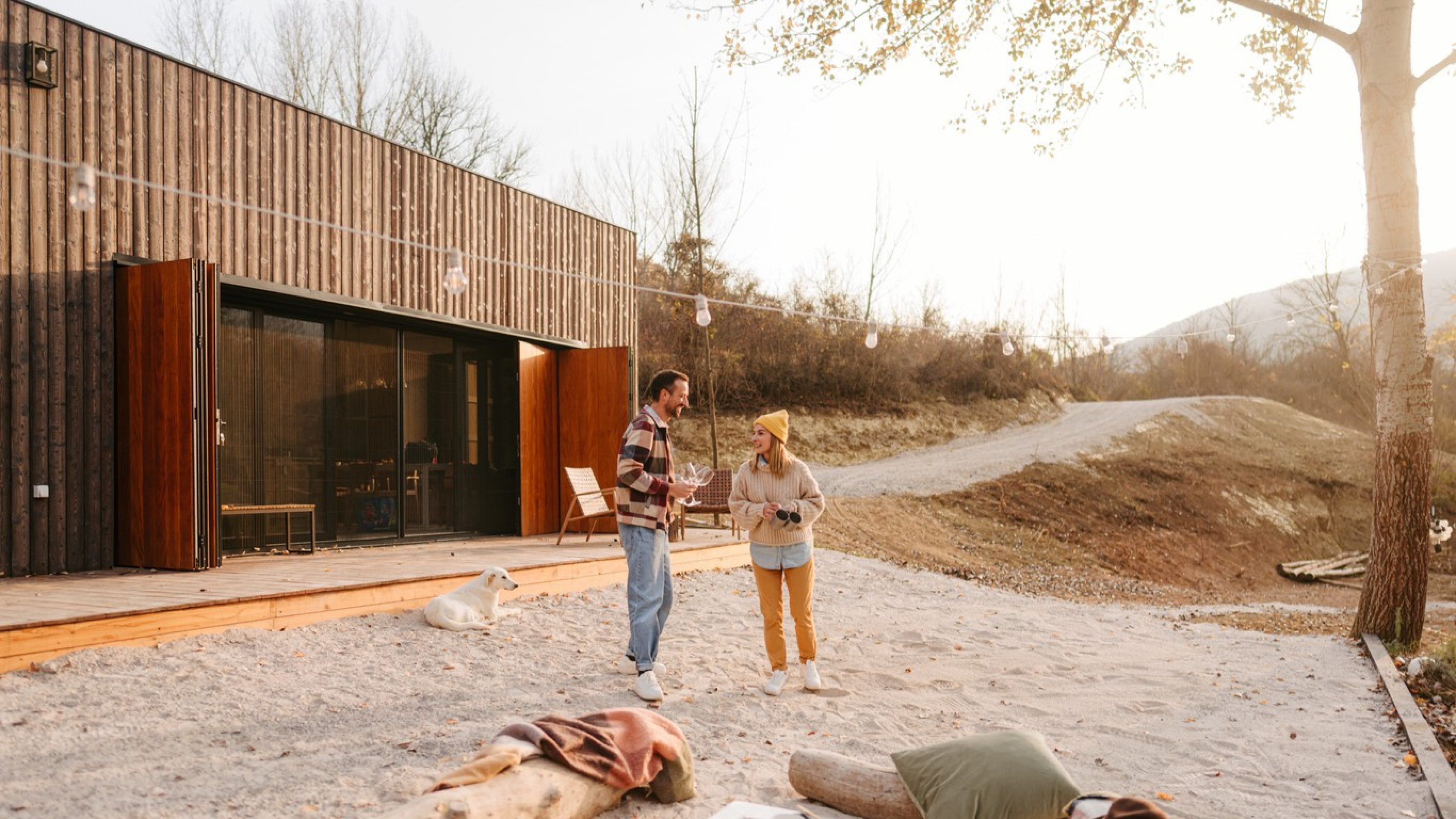 A happy couple is standing in the garden in front of a modern wooden house and talking. They’re dressed in casual clothes and are laughing. A dog is lying in the sand behind them, and in the background, you can see trees and hills. It is a sunny day and the picture conveys a relaxed, happy atmosphere.