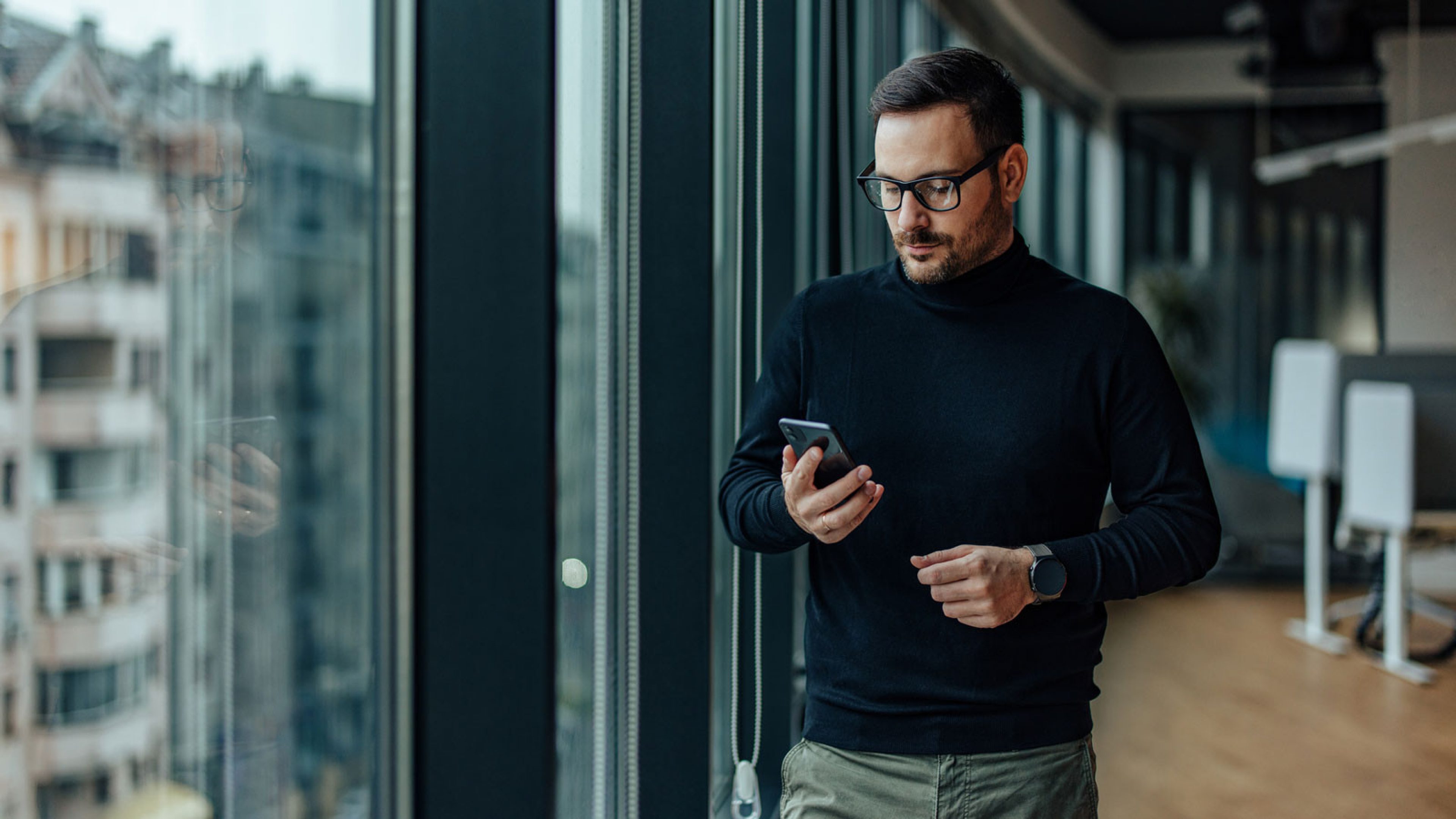 A middle-aged man stands next to a window façade with black frame elements. He is holding his mobile phone in his right hand. The man is in a modern residential building with elegant furniture.