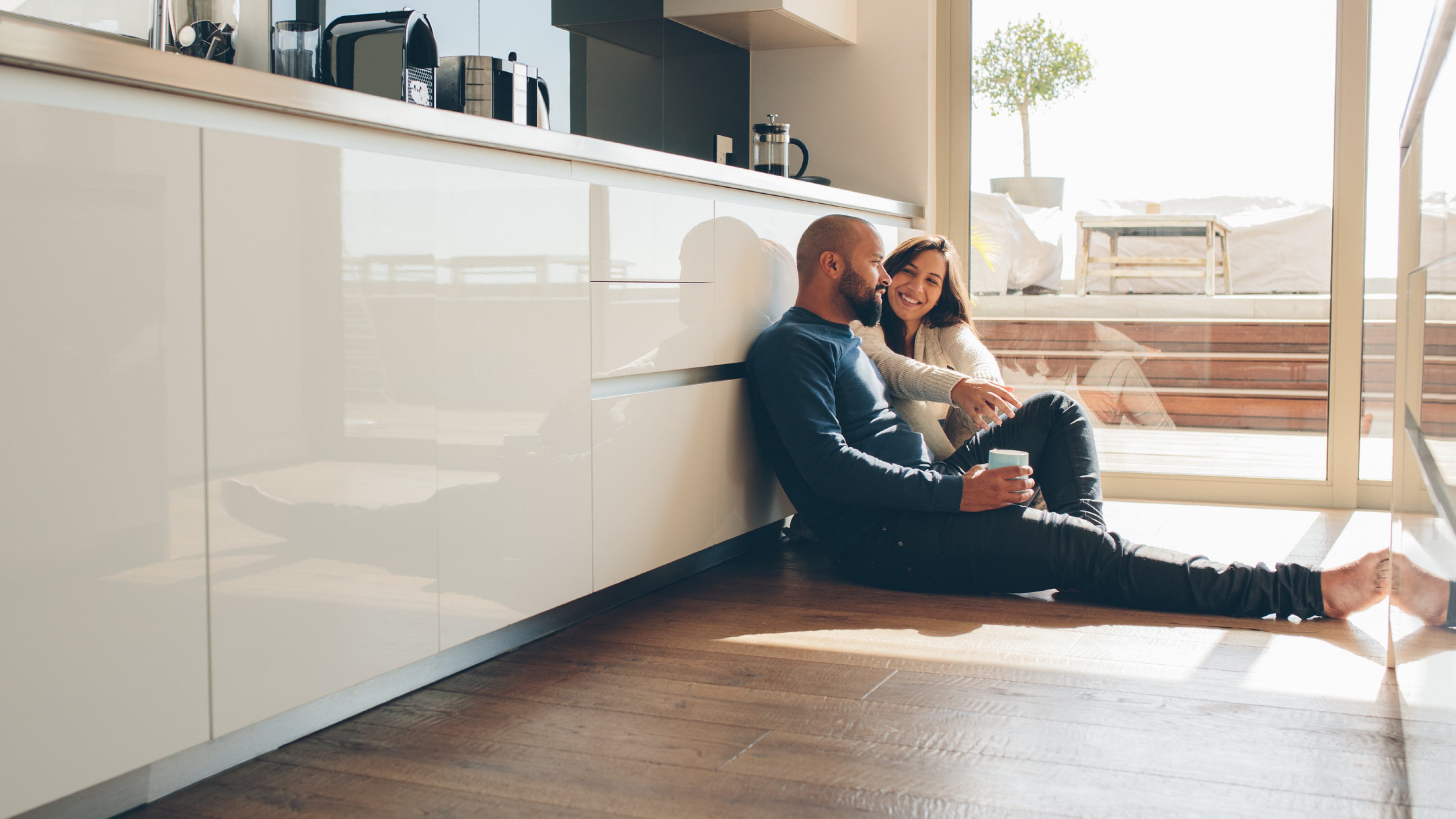 Two young people are sitting on the floor of a kitchen and talking. The man has a beard and a blue sweater, the woman is wearing a knitted sweater. Behind the couple you can see a roof terrace with covered patio furniture.