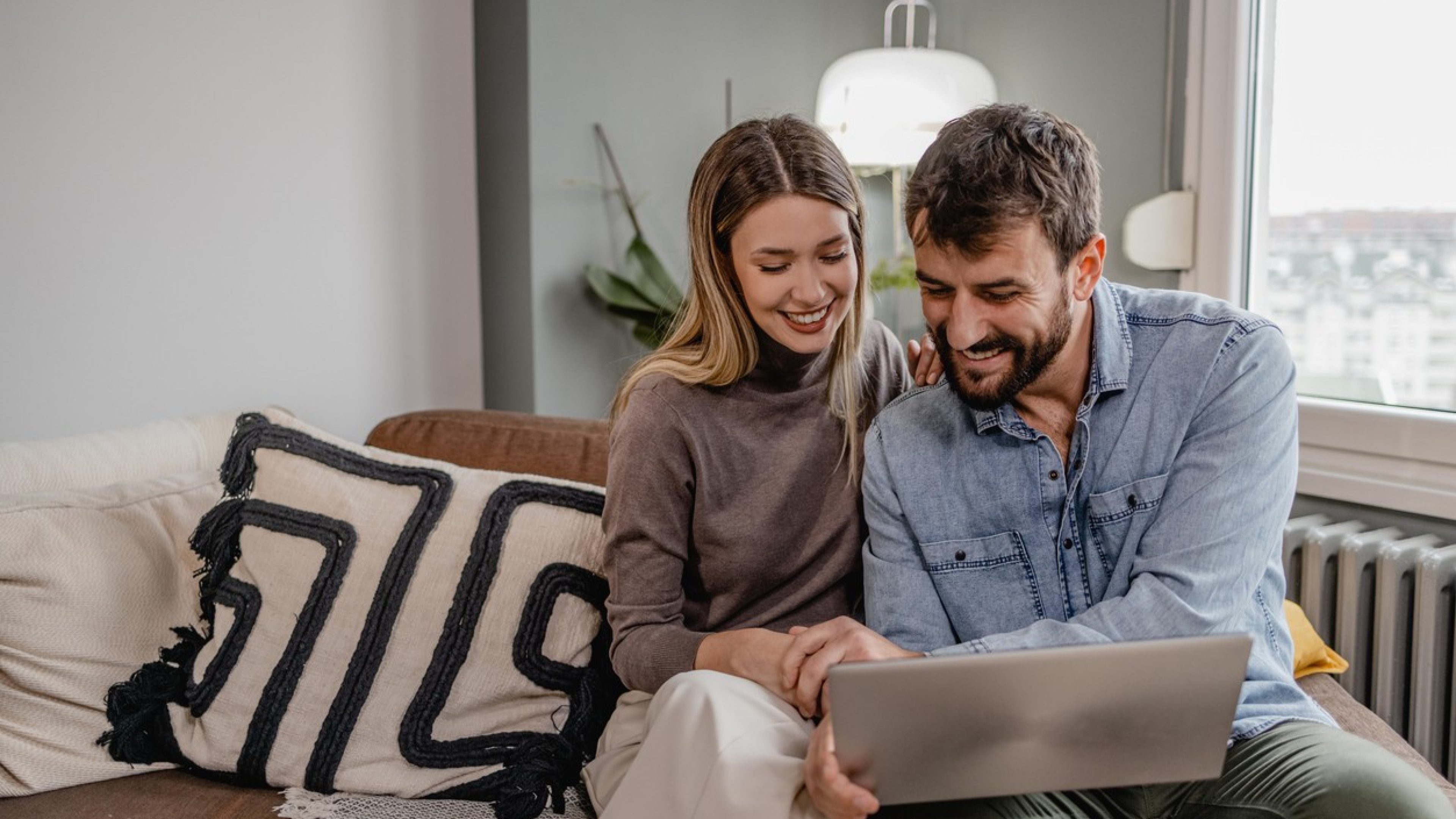A young couple is sitting on a sofa in a cosy apartment, smiling and looking at a laptop together. The woman has long blonde hair and is wearing a grey sweater, the man has dark hair and is wearing a light blue shirt. In the background are a modern decorative pillow, a plant and a lamp. The window in the background lets in natural light and shows an urban landscape. 