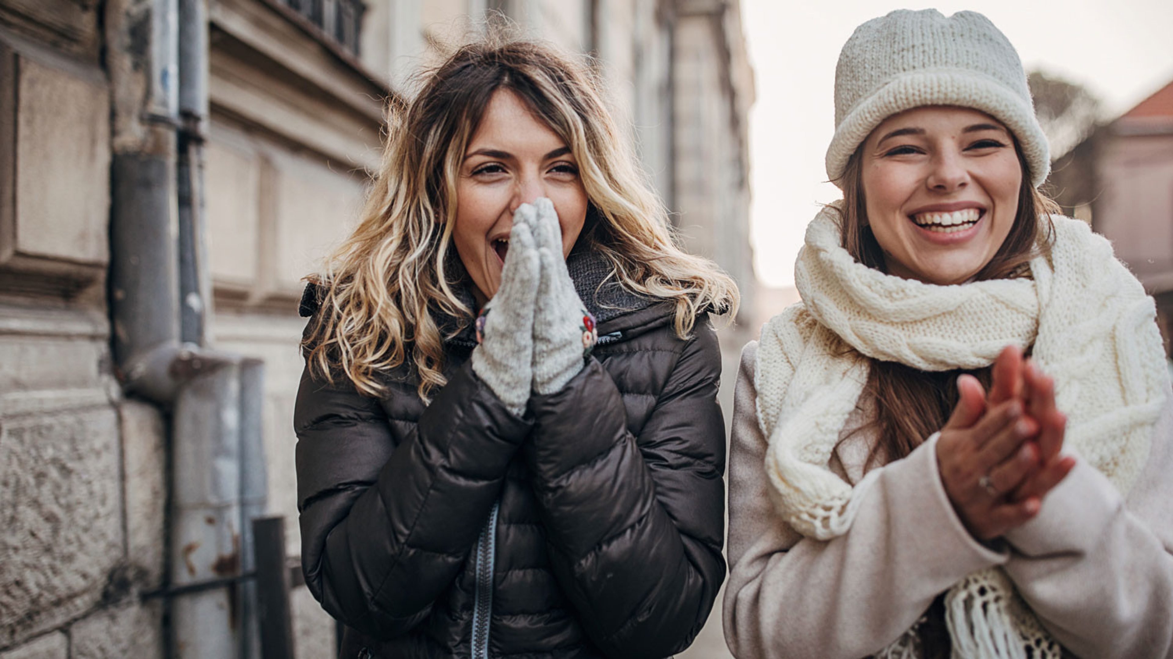 Two women are standing outside laughing. The woman on the left is blonde and is wearing a black down jacket and grey gloves with floral embroidery, the woman on the right is wearing a white cardigan, a white scarf and a matching white hat. Both women are laughing and happy.