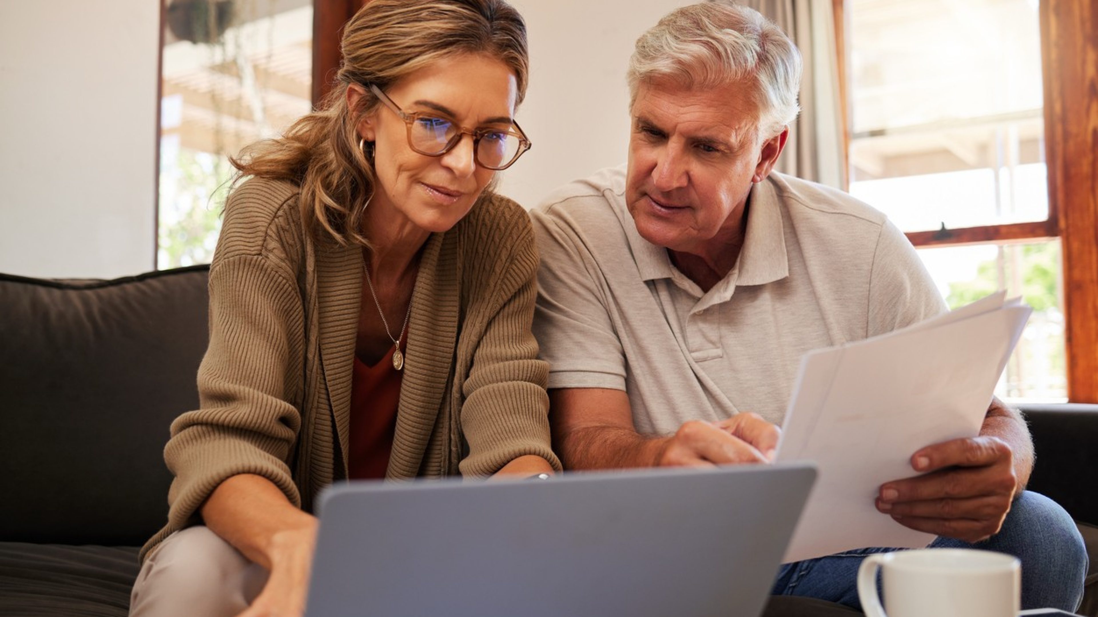 Two people, a woman with glasses and long hair and a man with white hair, are looking at documents. They are in a cozy, beautifully furnished living room.