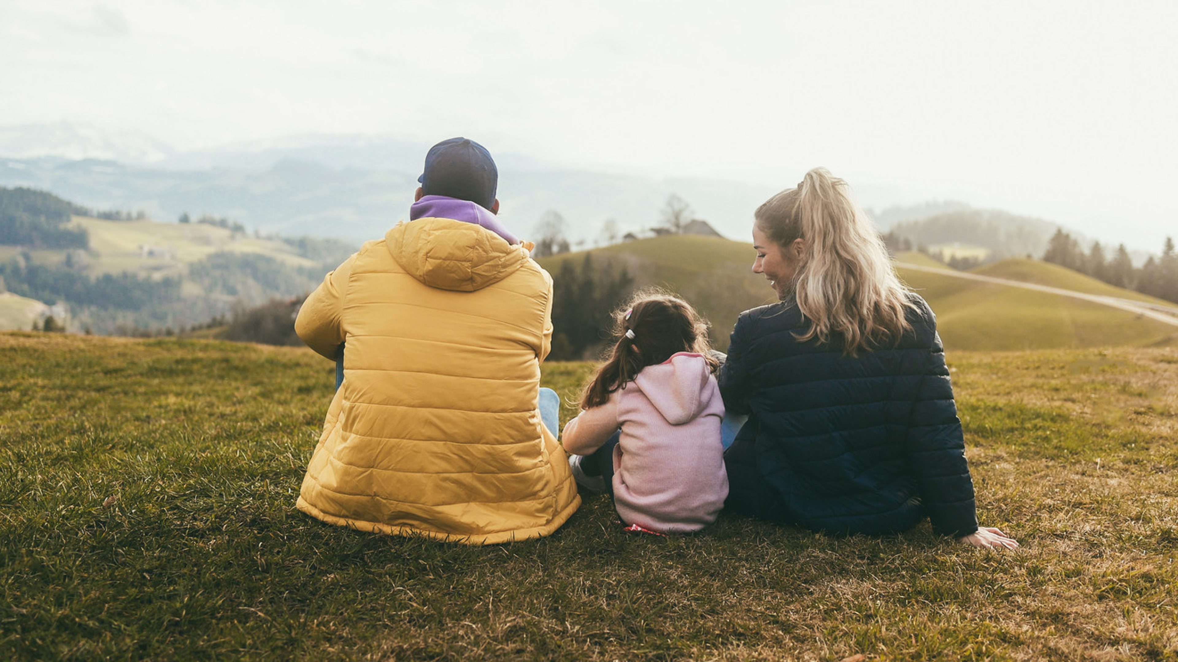 Two adults and one child are sitting with their backs facing on a meadow. They have a beautiful natural panorama in front of them. The man on the left is wearing a yellow down jacket, the child in the middle a pink sweater and the woman on the right a dark down jacket. She is looking at the child smiling. 