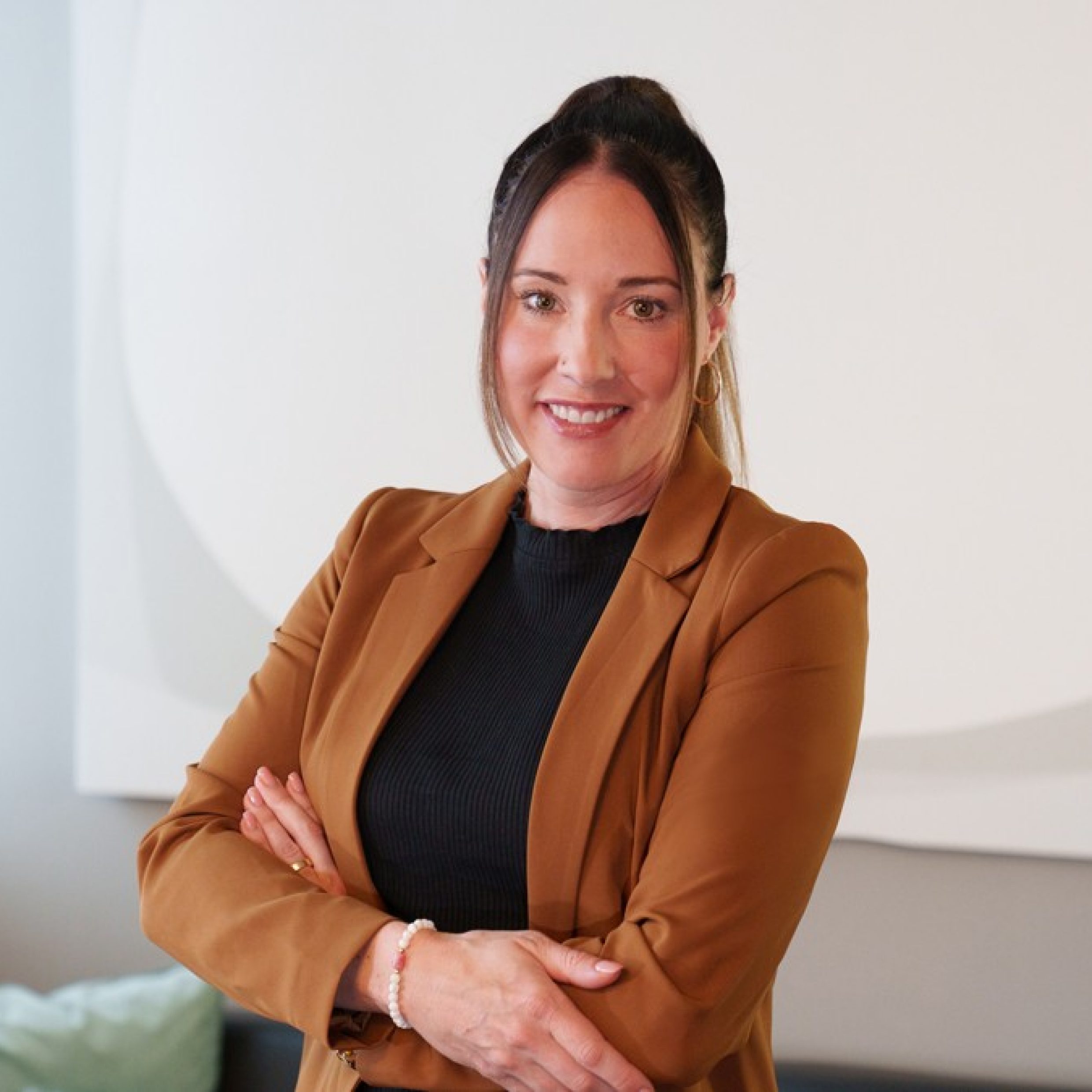 Portrait of a smiling woman wearing a brown blazer. The background features modern furniture and a cozy atmosphere.