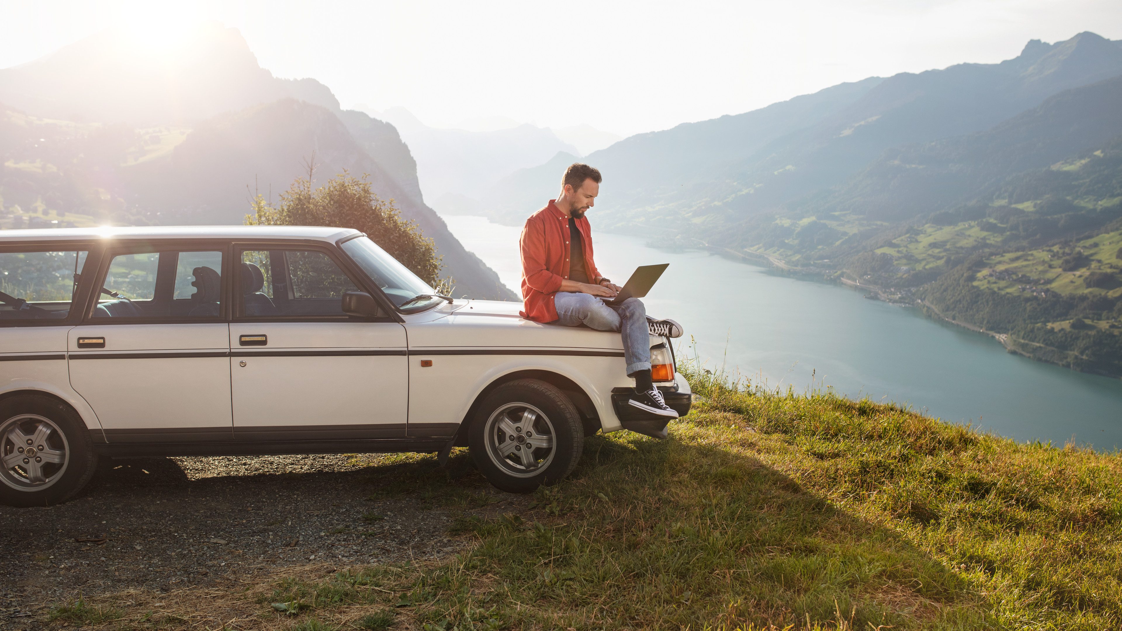 A young man is sitting on the bonnet of a white car reading something on his laptop. He is on a hill out in the countryside, behind him you can see a lake, mountains and sunlight. The man is wearing blue jeans, an unbuttoned red shirt and a dark T-shirt underneath.  