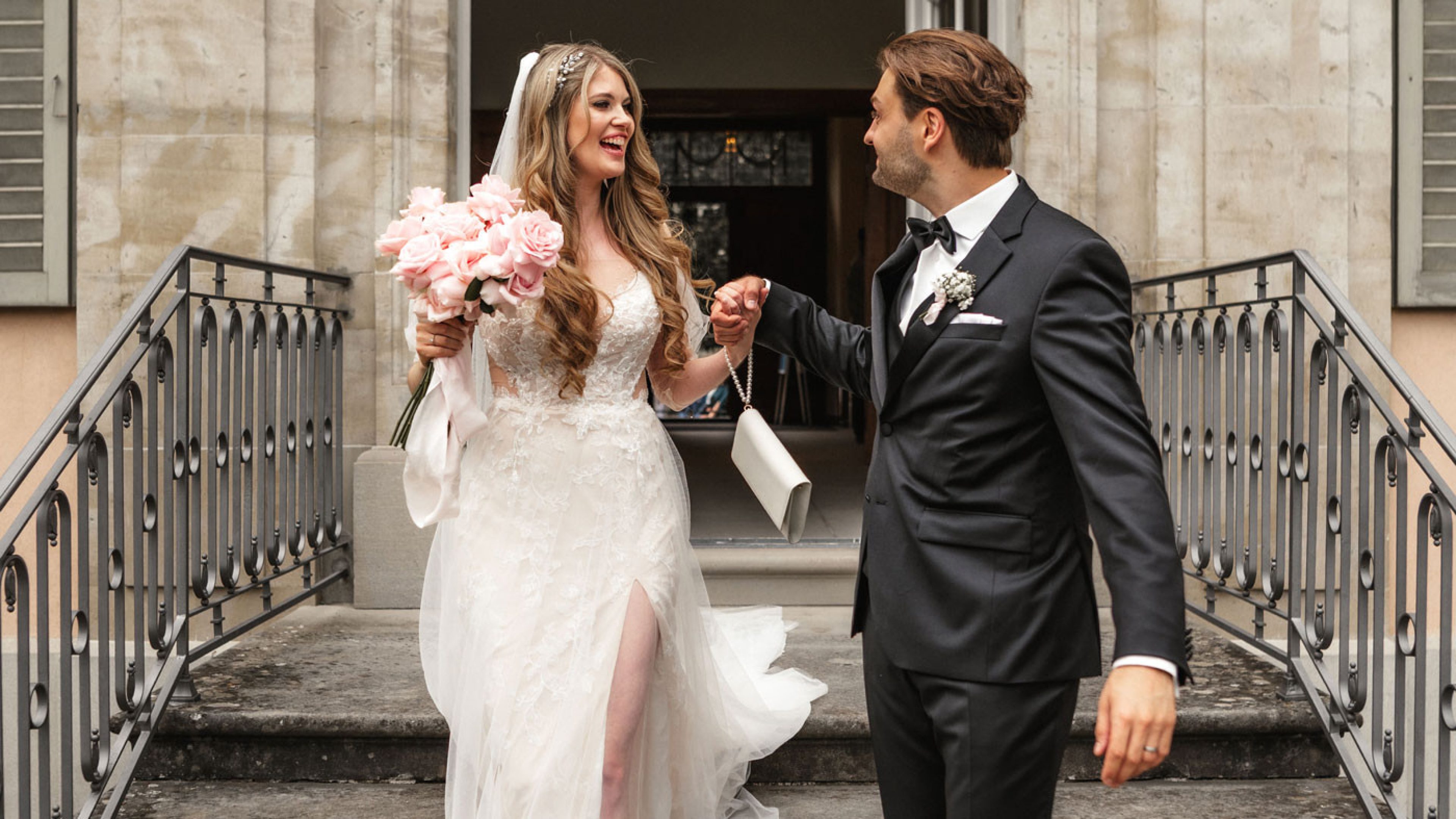 A newly married couple leaves the registry office via a stone staircase. The bride wears a white lace dress and holds a bouquet of pink roses in her hand. She has long, curly blonde hair with a white veil. They are both smiling at each other and holding hands as they walk down the stairs. The architecture of the building in the background looks classical with bright pillars and large windows.