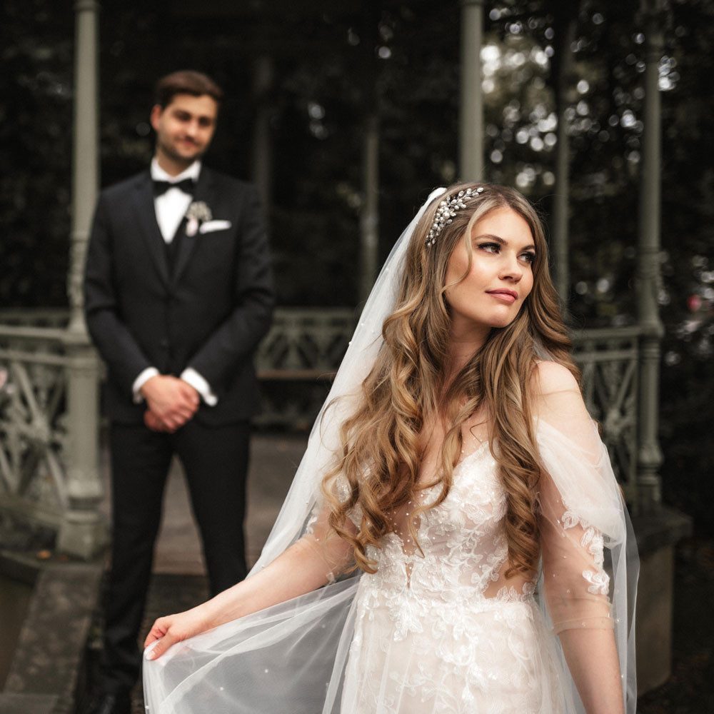 The bride stands in front of a pavilion, slightly turned to the side, with a dreamy look on her face. She is wearing an elegant dress with tulle sleeves and a hair trim. The groom stands in the background. He is wearing a suit and bow tie, his hands folded in front of his body. 