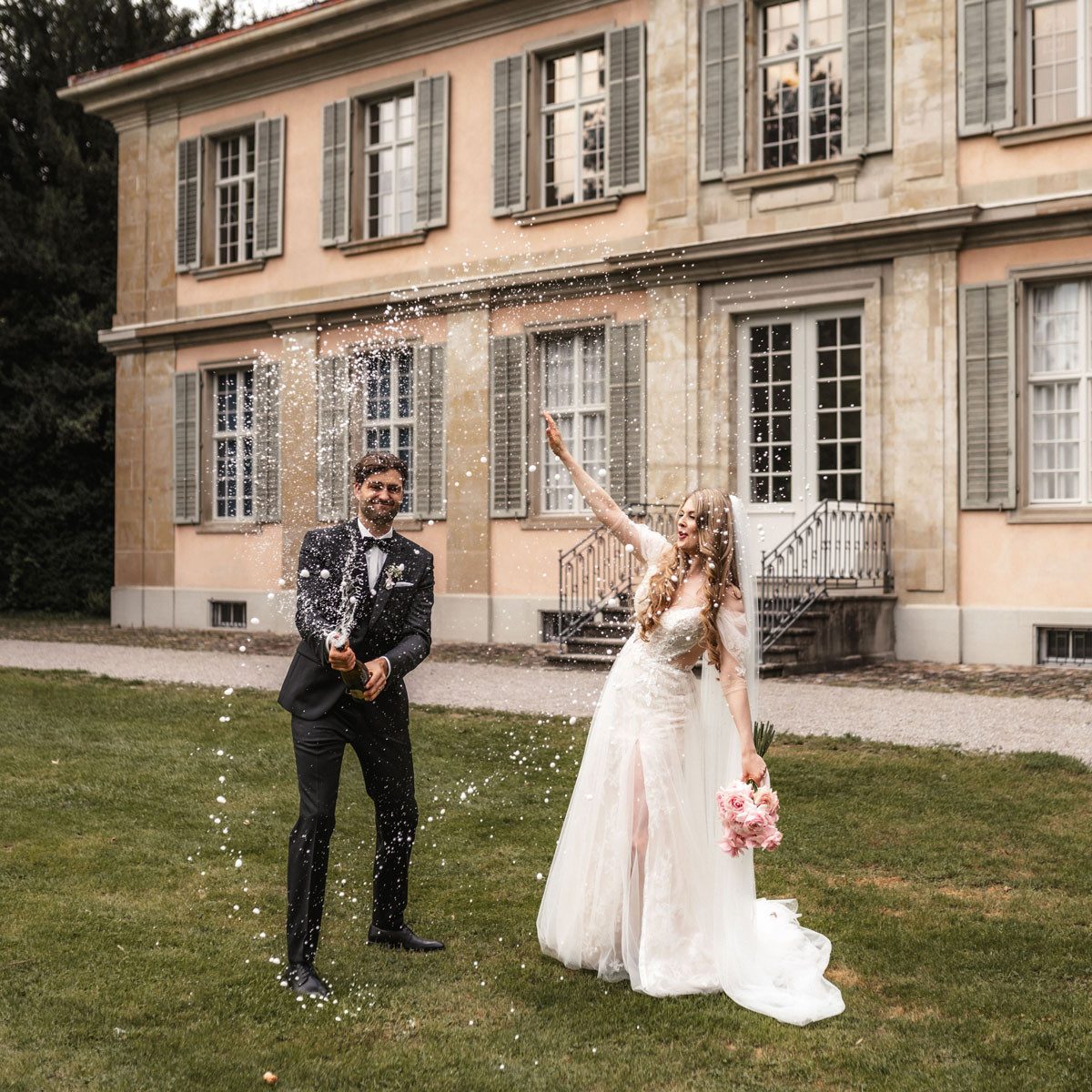 The bride and groom are standing in front of a villa laughing. The groom opens a champagne bottle, from which the foam splashes in a high arc. The bride stretches her arm into the air. The moment seems exuberant, cheerful and relaxed.