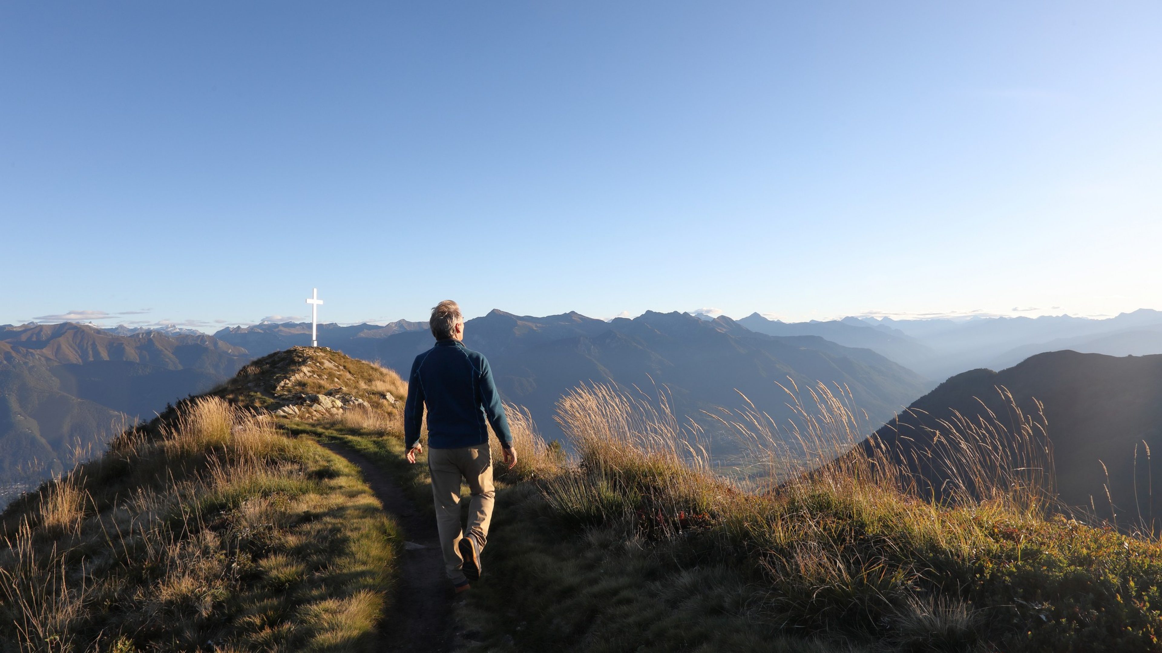 An older man walks on a grassy ridge at sunrise.
