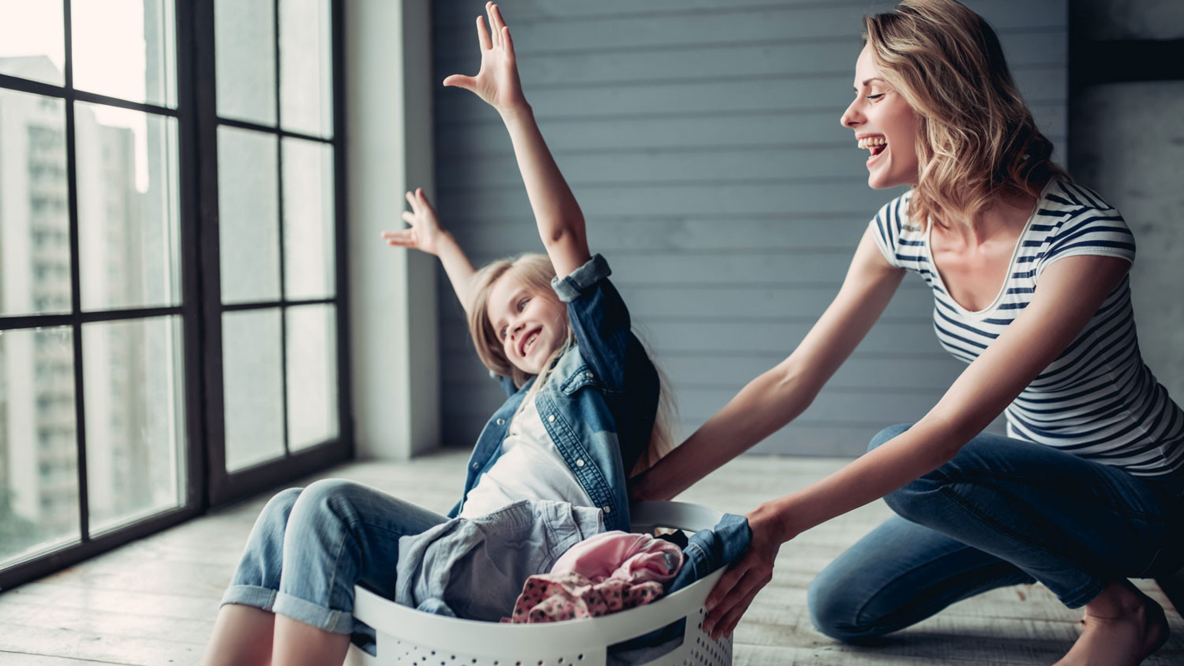 A young, blonde woman kneels on the floor and pushes her daughter towards the window in a laundry basket. The child waves her arms in the air and enjoys the “train” game.