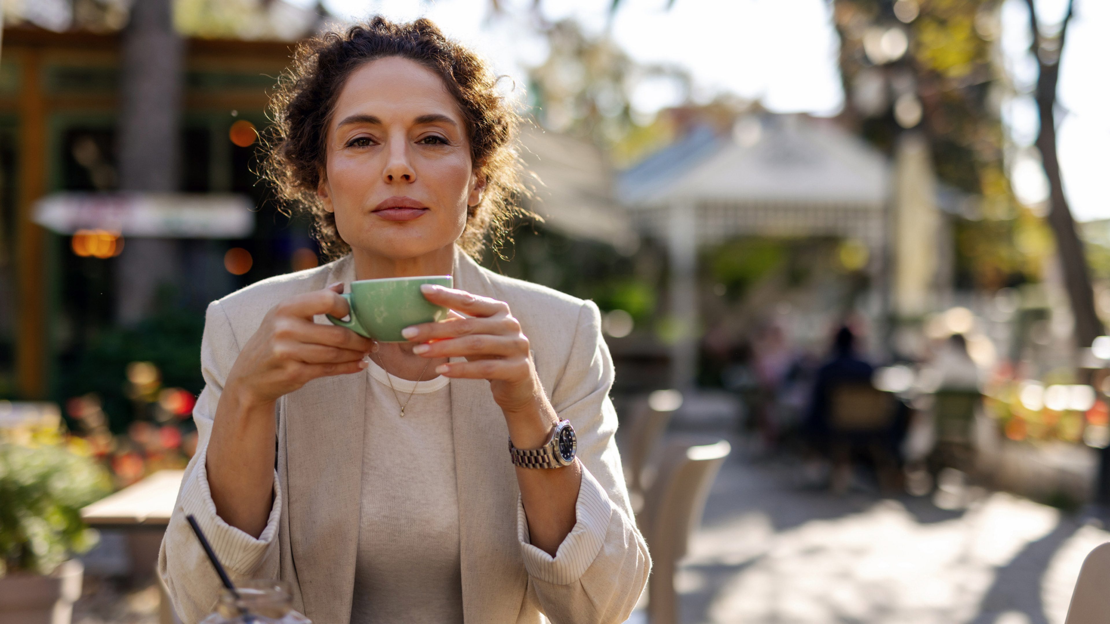 A woman is sitting relaxed in a sunny outdoor café. She is holding a green coffee cup with both hands and looking calmly at the camera. She is wearing a bright outfit with a blazer and watch. Other customers can be seen in the background.