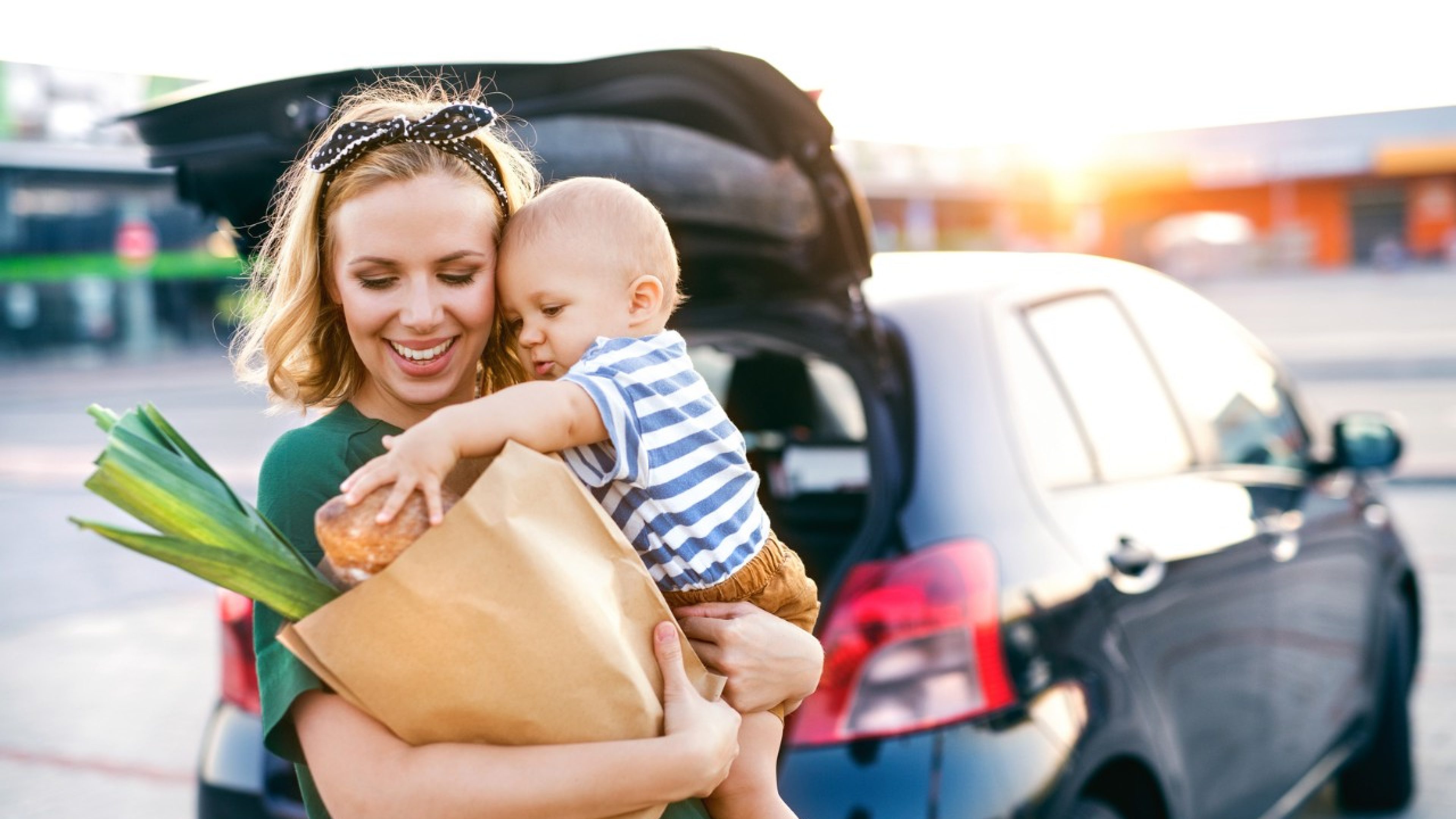 Beautiful young mother with her little baby son in front of a supermarket, holding paper shopping bag. Woman with a boy standing by the car.