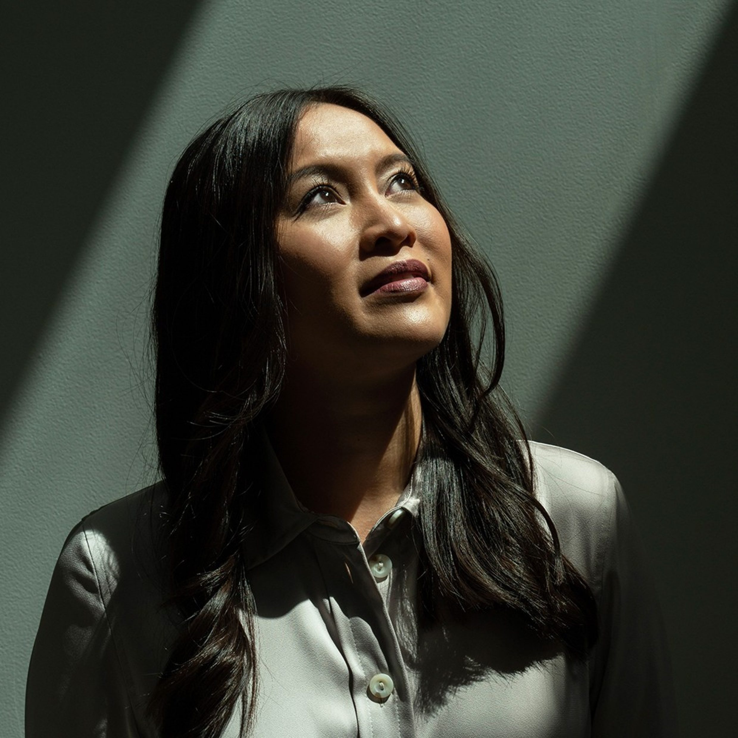 A black-haired woman in an elegant white blouse looks up at a ray of light falling on her.