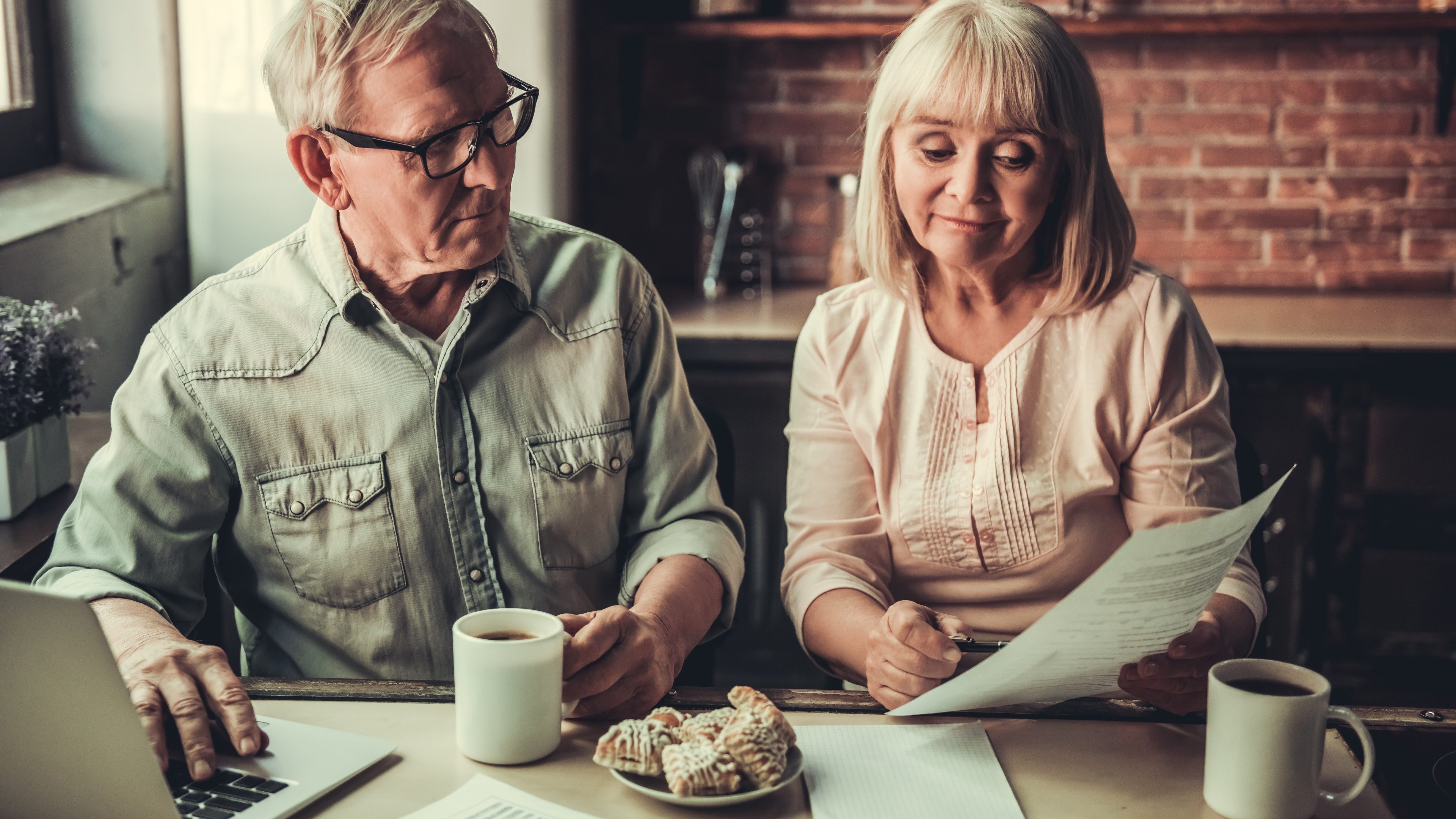 Beautiful senior business couple is drinking tea, eating cookies, using a laptop and smiling while working in kitchen