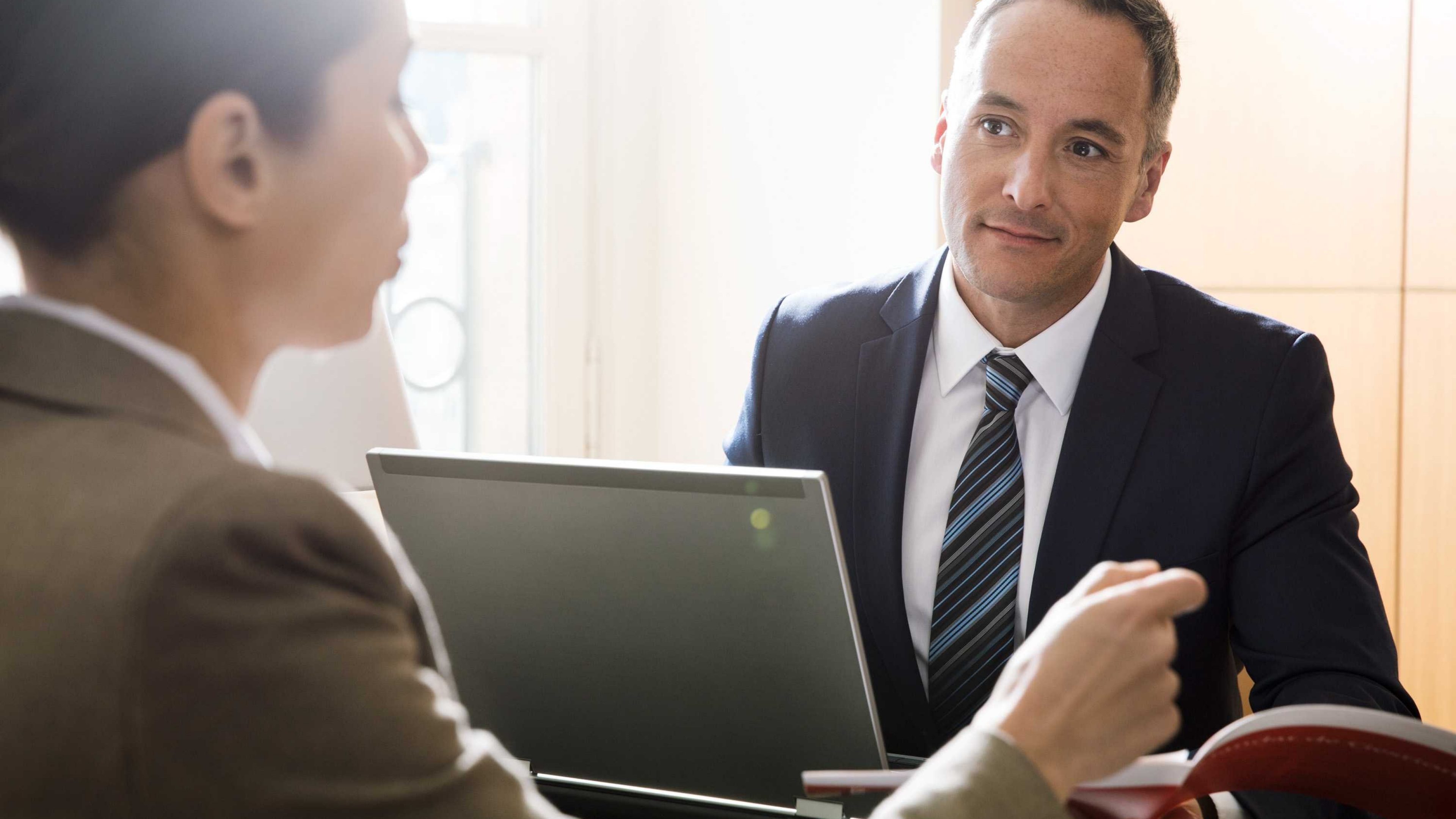 Two employees are discussing a business case in the office. A laptop is on the desk and specialist literature is open. 