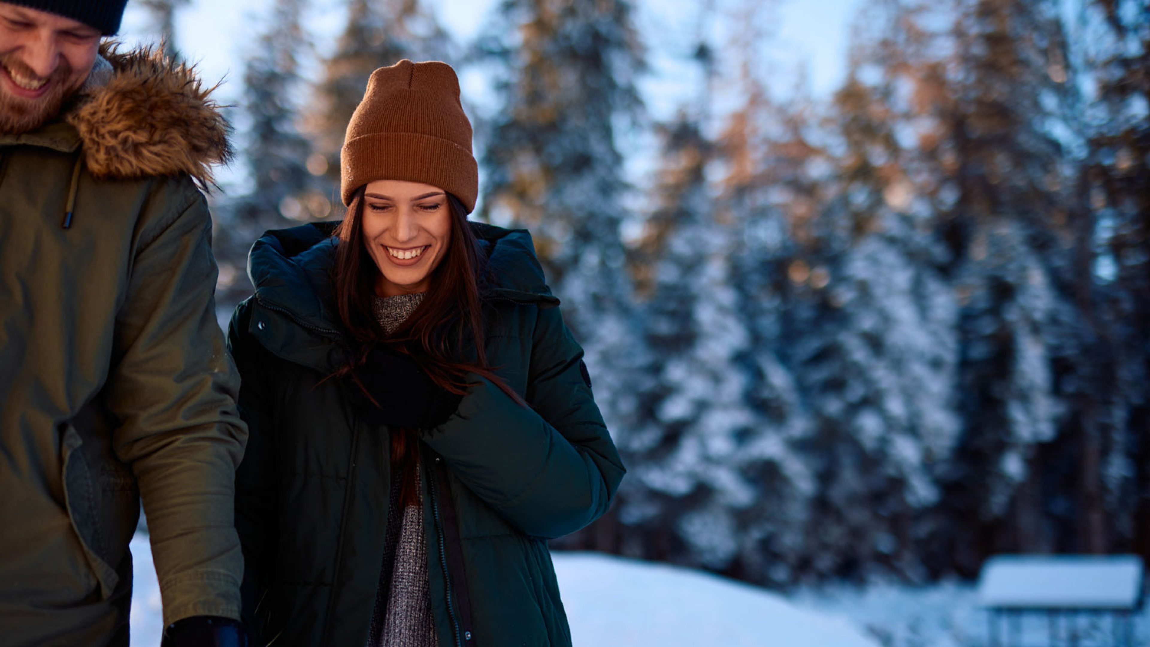 Two women are standing outside laughing. The woman on the left is blonde and is wearing a black down jacket and grey gloves with floral embroidery, the woman on the right is wearing a white cardigan, a white scarf and a matching white hat. Both women are laughing and happy.