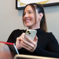 A woman is sitting on a chair, relaxed and smiling, and holding a smartphone in her hand. She is wearing a black, long-sleeved top and has a notepad on her lap. There is an abstract painting on the wall behind her.