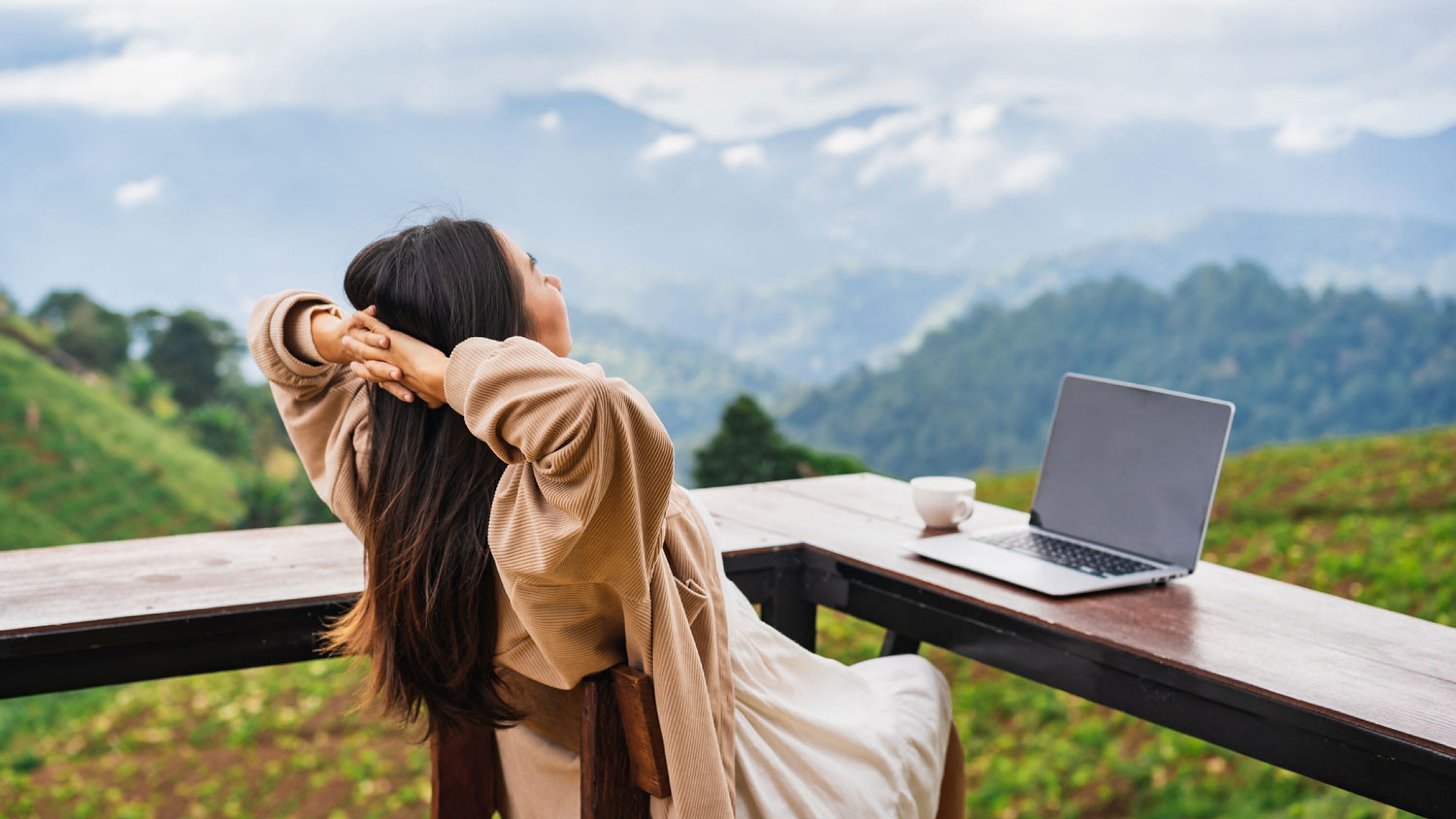 A person is sitting at an outdoor table with a laptop and a cup of coffee, stretching with their hands behind their head and looking out at a mountainous, green landscape