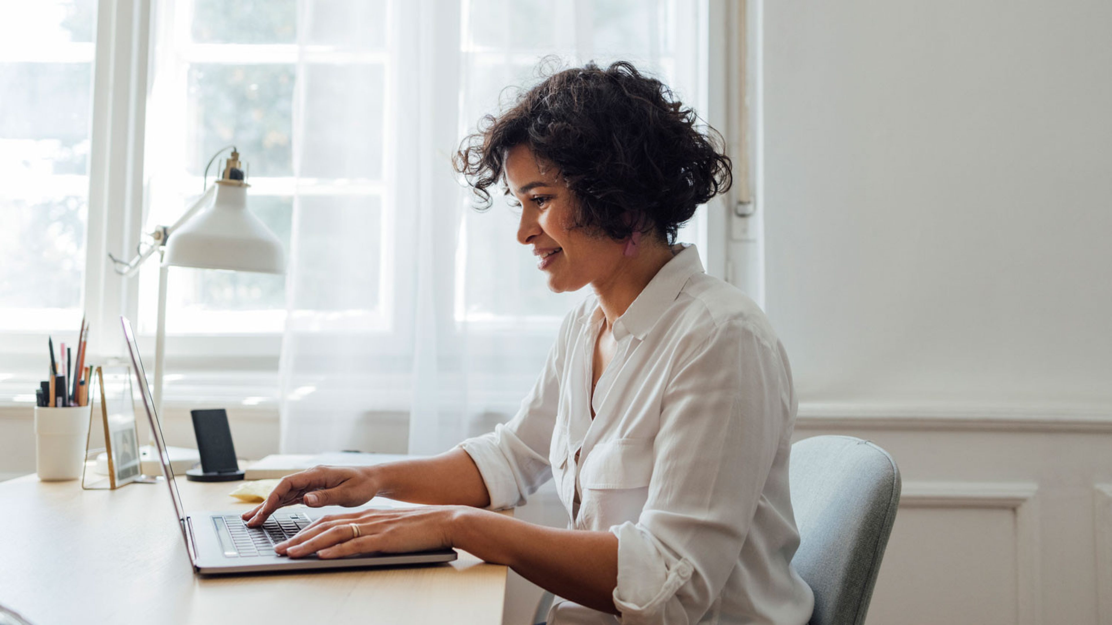 A woman with dark, curly hair is sitting at a desk and working on a laptop. She is wearing a white, long-sleeved shirt with an open collar. The room is bright and welcoming, with large windows and white curtains in the background. On the desk, there is a modern desk lamp, a smartphone, a notebook, and a few pens.