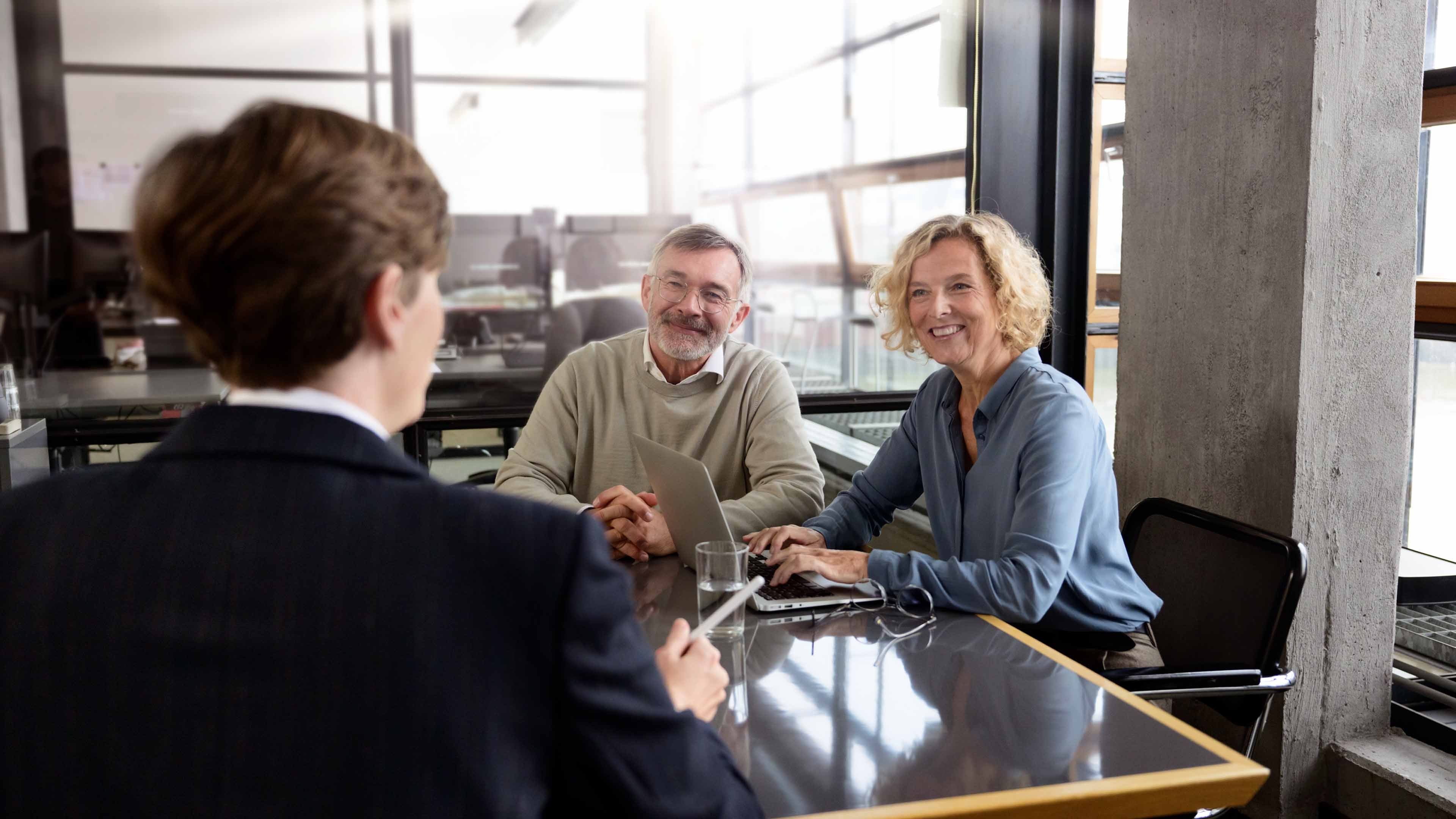 Three people in a modern office setting during a business meeting. Two are facing the camera, smiling, while one is seen from behind. A laptop and office equipment are visible.