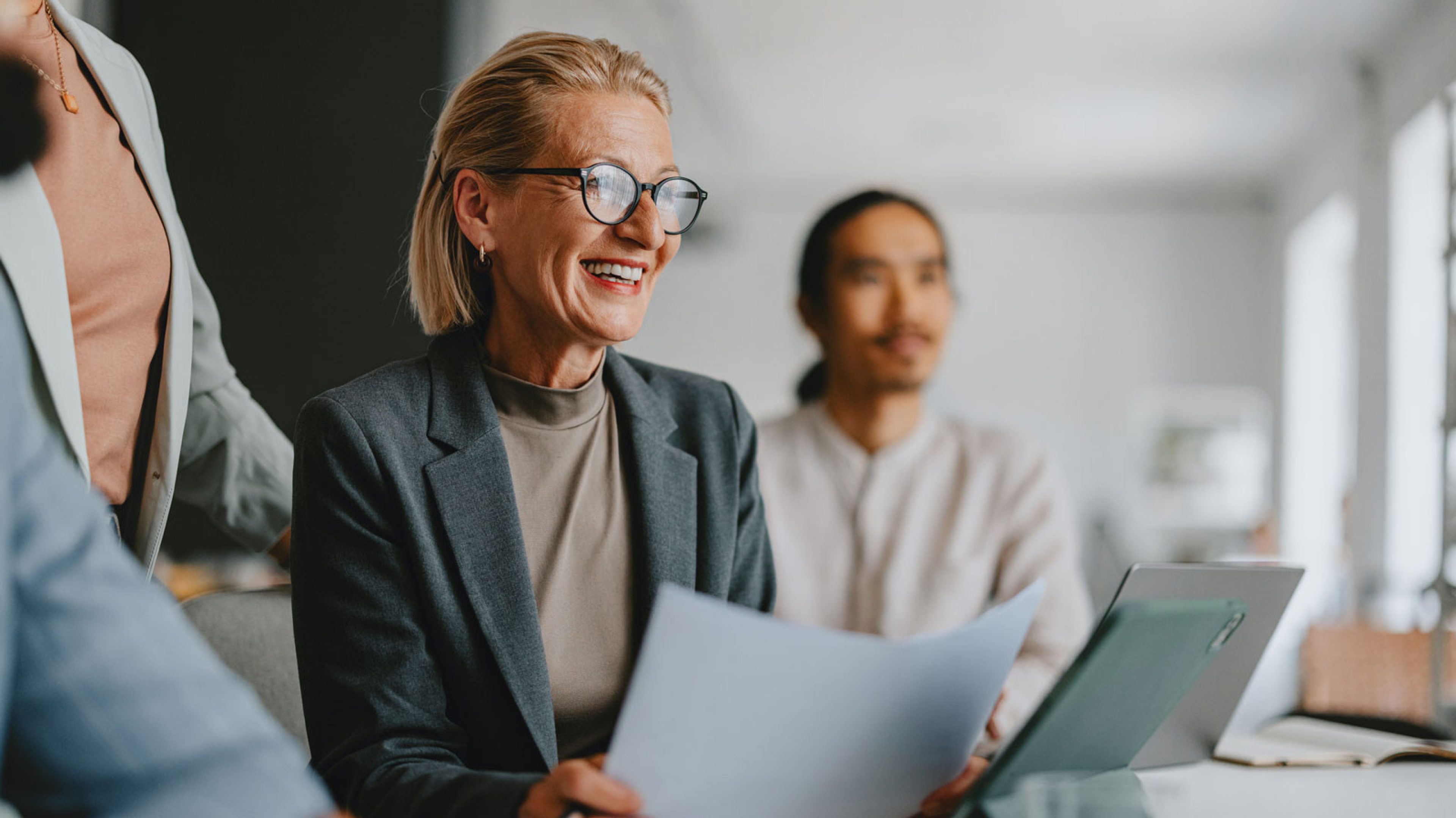 A well-dressed businesswoman in her mid-fifties smiles and holds documents in a meeting while a colleague sits in the background. In front of her is a tablet. The scene conveys an atmosphere of collaboration and professionalism.