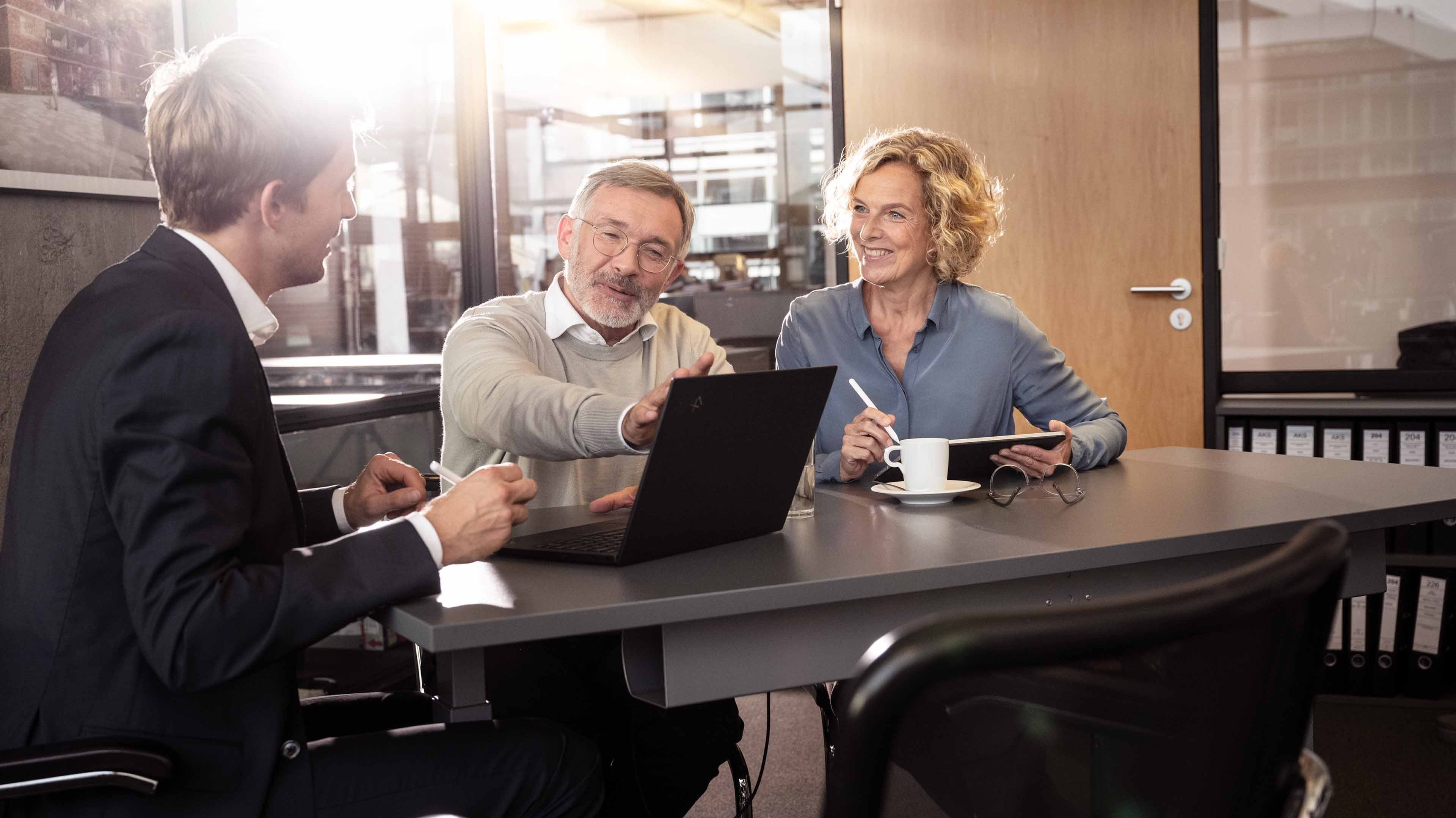 A consultant sits with two entrepreneurs at a table in an office, pointing at a laptop. The entrepreneurs listen attentively and take notes.