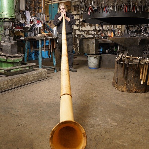 A man stands in a blacksmith’s shop playing an alphorn.