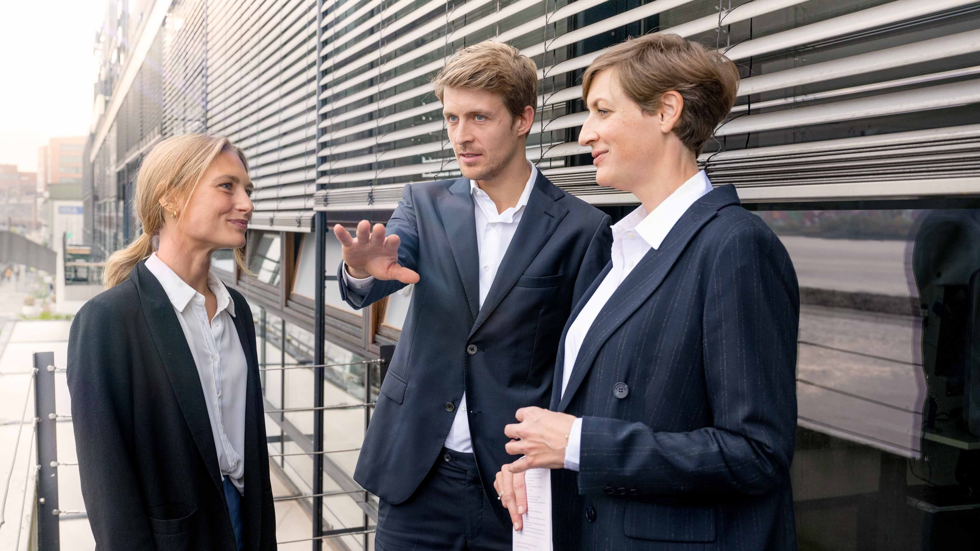 Two women and a man in black blazers in front of a company building