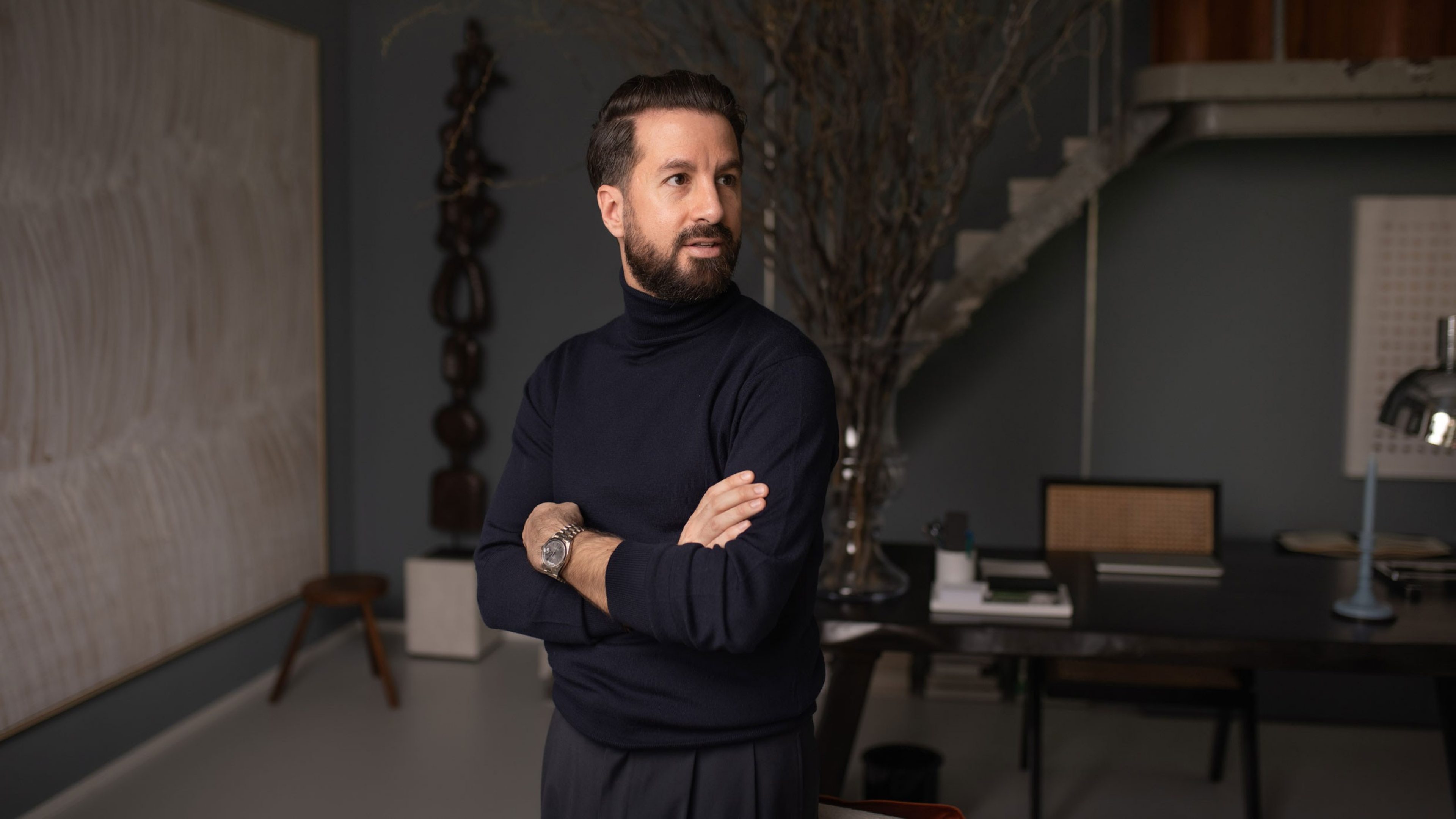 Un homme vêtu d’un pull à col roulé de couleur sombre se tient debout, les bras croisés, dans un bureau élégamment meublé. Il porte une montre et regarde de côté, l’air pensif. A l’arrière-plan se trouve un grand bureau avec des accessoires minimalistes. Un escalier mène à l’étage, avec à son côté une œuvre d’art abstrait et une grande branche sèche dans un vase qui servent de décoration. L’ambiance est moderne et professionnelle.