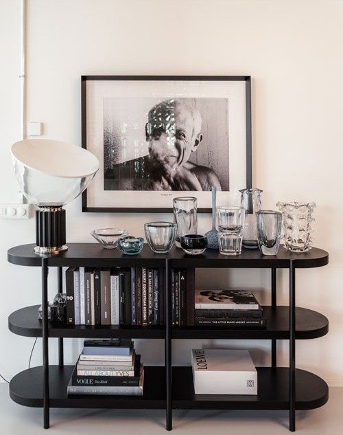 Une étagère noire, soigneusement garnie de livres, d’élégants accessoires pour la maison et d’une sélection d’objets en verre. Au-dessus de l’étagère, une photo en noir et blanc d’un homme est suspendue. Une lampe élégante complète la décoration sophistiquée.