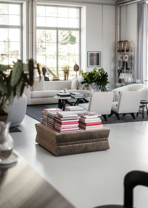 Un coup d’œil dans un salon élégant, au centre duquel se trouve une pile de livres posés avec style sur un pouf rembourré. L’arrière-plan est dominé par un élégant ensemble de fauteuils et canapés clair installé dans un coin avec de grandes fenêtres à travers lesquelles jaillit une lumière naturelle. 
