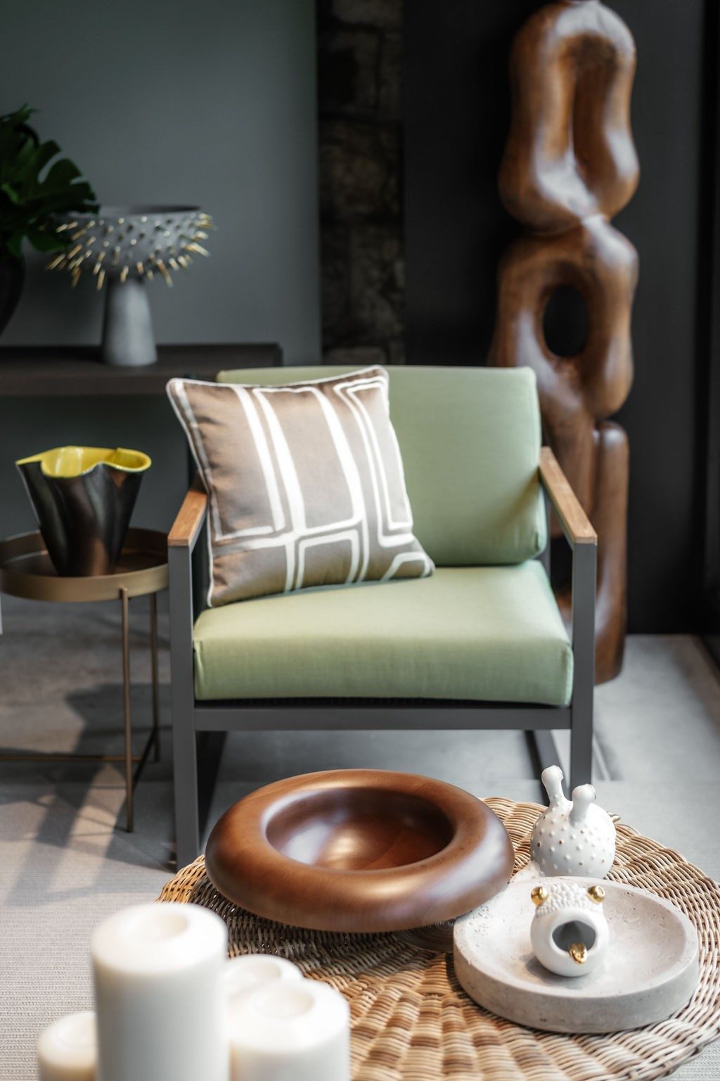 Un fauteuil vert, sur lequel est déposé un coussin, se trouve derrière une table basse ronde en bois tressé. Sur la table sont posés des plats creux et des articles de décoration. L’un des articles de décoration a la forme d’une tête de grenouille aux yeux dorés qui tire une langue dorée. Derrière le fauteuil se trouvent deux objets: une sculpture en bois en forme de chaîne et un récipient avec des éléments pointus.