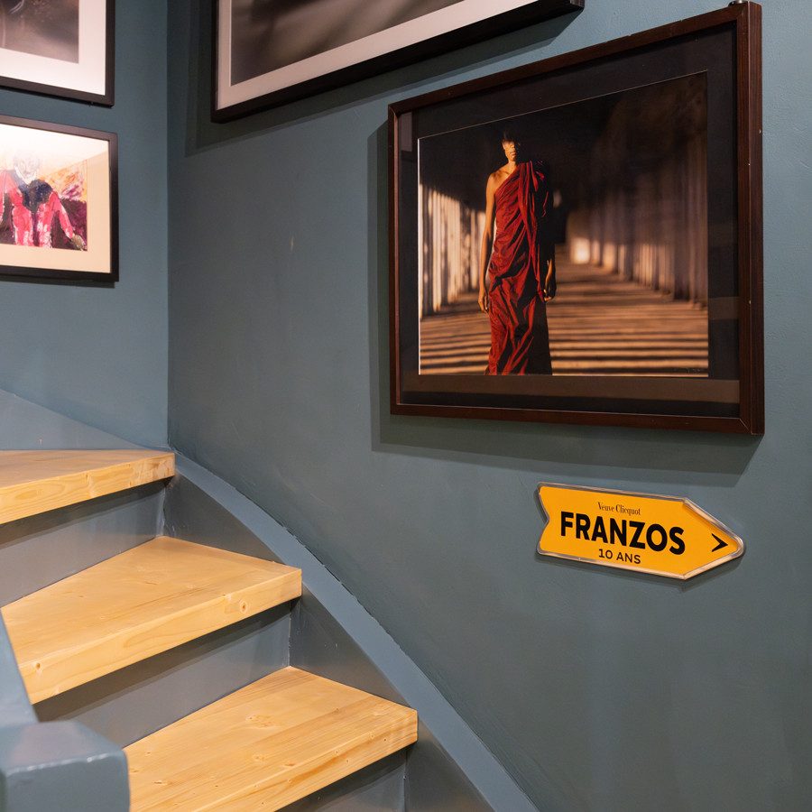 Un escalier avec des marches en bois clair qui mène en haut vers la gauche. Le mur le long de l’escalier est peint en bleu et en gris, il est décoré avec plusieurs photos encadrées. Au centre, on remarque une grande photo montrant une personne vue de derrière, vêtue d’un vêtement rouge et traversant un couloir bordé de piliers. Au-dessous de cette image se trouve un panneau jaune sur lequel figurent les lettres «FRANZOS» en noir et une flèche vers la droite. En dessous, il est écrit «50 MINS » en caractères plus petits.