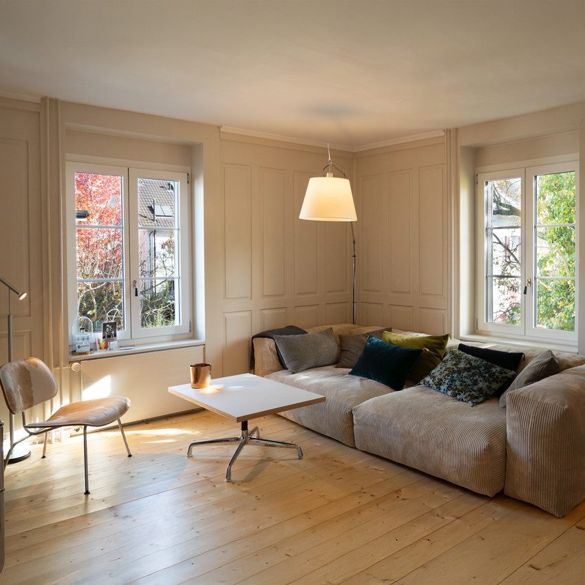 Une pièce lumineuse avec un parquet et un grand canapé gris. Plusieurs coussins de couleurs différentes sont posés sur le canapé. Devant celui-ci se trouvent une petite table rectangulaire dotée d’un plateau blanc et d’un piètement en métal; une chaise avec une assise claire est visible à côté. Les murs sont recouverts de lambris et deux fenêtres laissent pénétrer la lumière naturelle de chaque côté. 