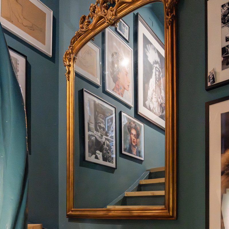L’illustration montre un grand miroir doré avec un cadre décoré accroché à un mur bleu et vert. Dans le miroir, on peut voir un escalier avec des marches en bois clair qui mène vers le haut. Plusieurs photos encadrées, de différents formats et tailles, sont accrochées le long de l’escalier. Le mur sur lequel est posé le miroir est également orné de photos encadrées.