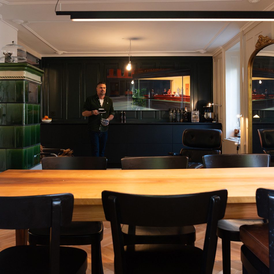 L’illustration montre une pièce avec une grande table en bois visible au premier plan, entourée de plusieurs chaises noires. A l’arrière-plan se trouve une kitchenette sombre avec des armoires et un plan de travail. Sur l’image à gauche, on voit un grand poêle en faïence vert. Un grand miroir avec un cadre doré est accroché au mur à droite. Un homme traverse la pièce et porte une tasse de café noire.