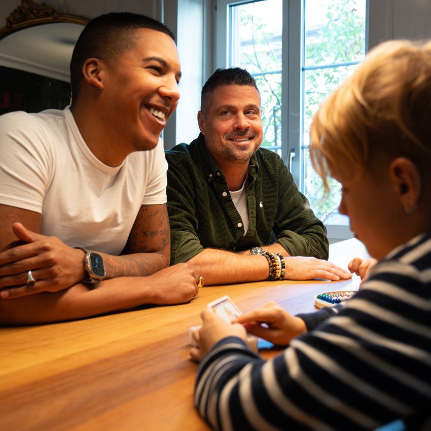 Deux adultes sont assis à une table en bois et ont les bras posés sur la table. Un enfant est assis en face et tient des cartes dans une main. En arrière-plan, on peut voir de grandes fenêtres à travers lesquelles des feuilles vertes sont visibles.