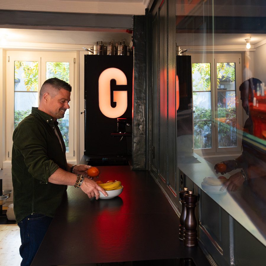 La photo montre une cuisine moderne et ouverte, avec de grandes fenêtres et beaucoup de lumière naturelle. Une personne se tient devant un long plan de travail sombre et prépare des fruits frais. 