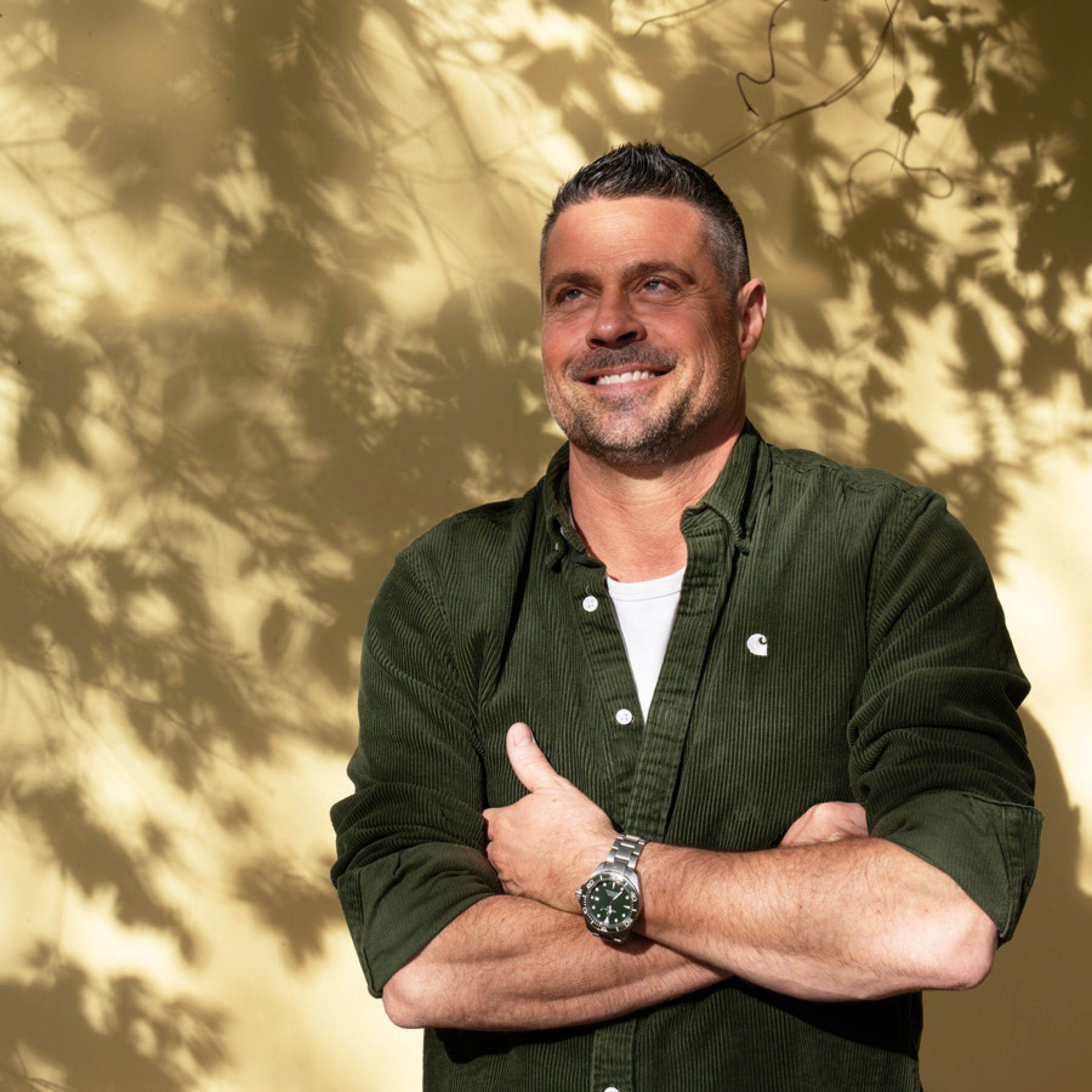 L’illustration montre un homme se tenant debout devant un mur jaune pâle. Sur le mur, on distingue des ombres de feuilles et de branches qui forment un motif irrégulier. L’homme porte une chemise vert foncé avec des boutons et un t-shirt blanc en dessous. L’homme a les bras croisés devant le corps.