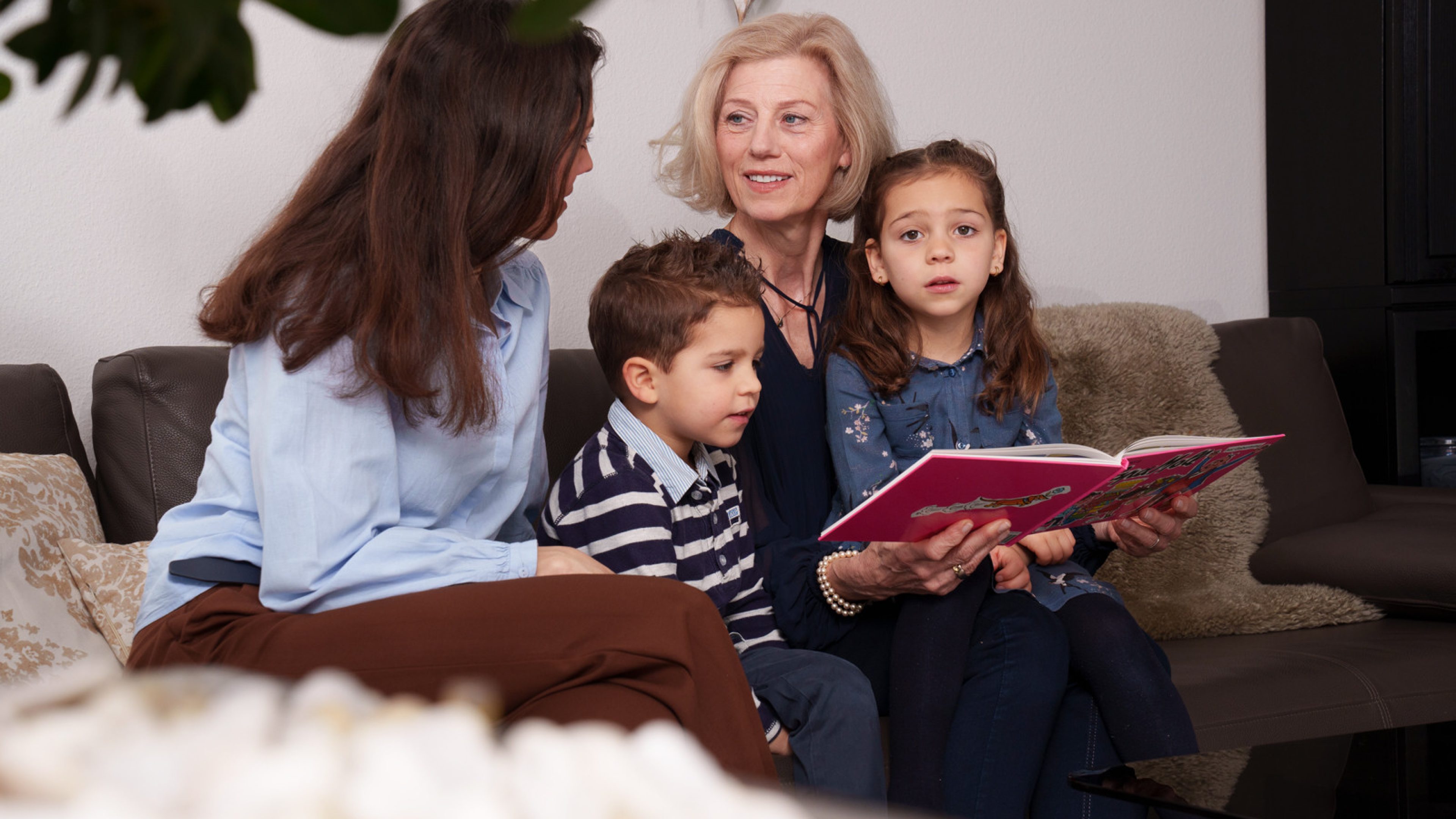 Une belle femme aux cheveux coupés courts est assise avec sa fille et ses petits-enfants sur un canapé marron foncé avec des coussins et des couvertures beiges et marron. Elle tient un livre rose; une jeune fille aux cheveux bruns qui regarde timidement la caméra est assise sur ses genoux. A côté, un petit garçon vêtu d’un pull avec des rayures bleues et blanches regarde le livre. Une jeune femme vêtue d’un chemisier bleu clair et d’un pantalon marron est assise à côté de lui. Elle discute avec la femme aux cheveux coupés courts.