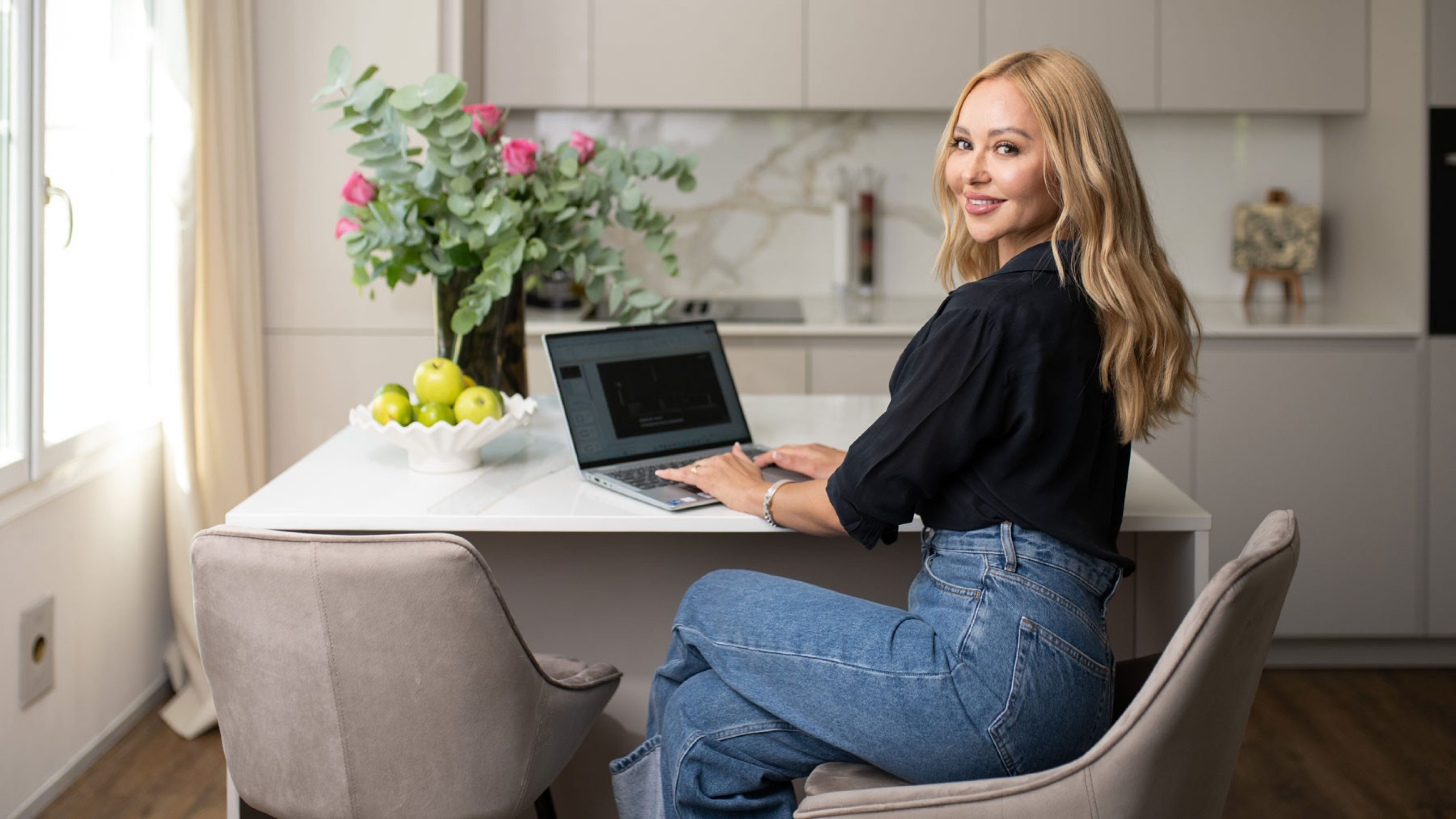 Une femme blonde est assise à un îlot de cuisine sur un tabouret de bar. Elle sourit et regarde l’objectif par-dessus son épaule. En face d’elle est ouvert un ordinateur portable, à côté duquel se trouve un magnifique bouquet de fleurs.