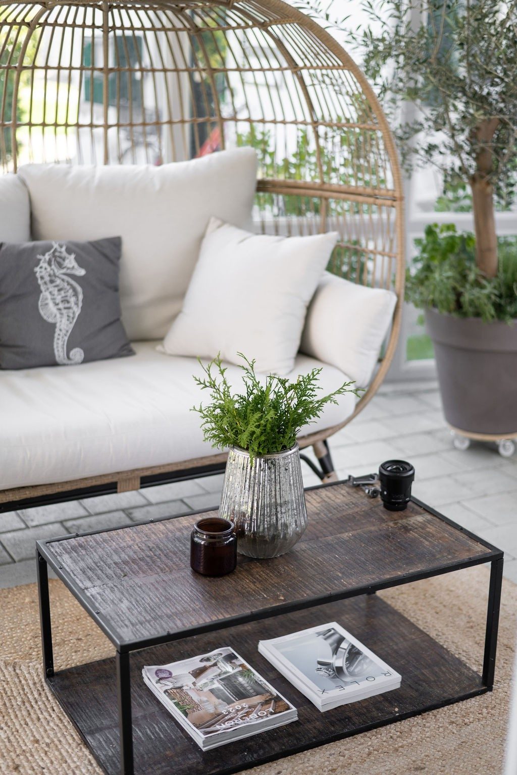 Devant un canapé lounge en osier se trouve une table d’appoint en bois sombre ornée d’une plante verte et de magazines bien rangés.