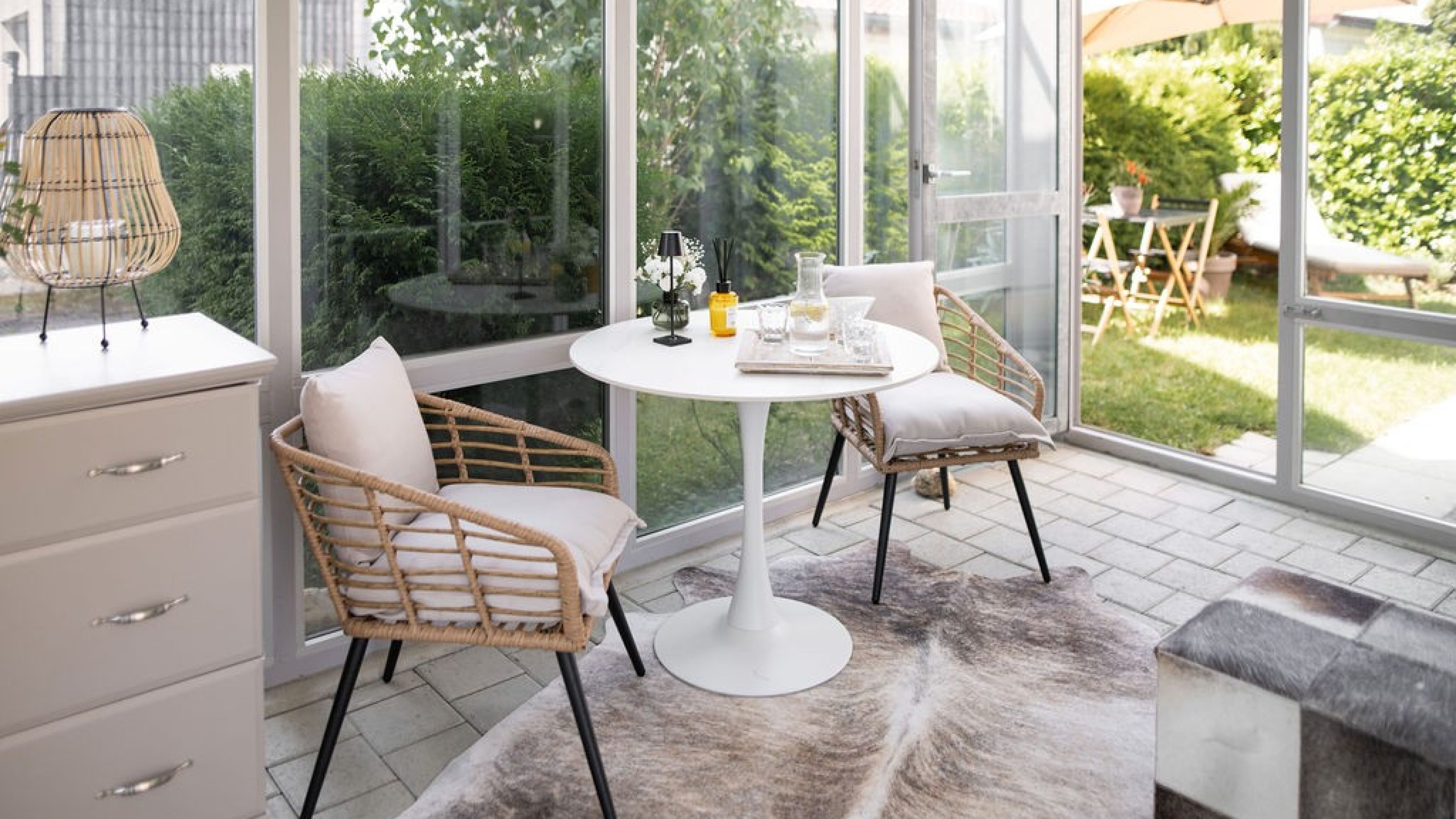 Dans un jardin d’hiver, une petite table ronde et deux chaises ont été installées sur une peau d’animal. Sur la table se trouvent des verres à eau et une carafe à eau, ainsi qu’une lampe et un diffuseur de parfum d’intérieur.