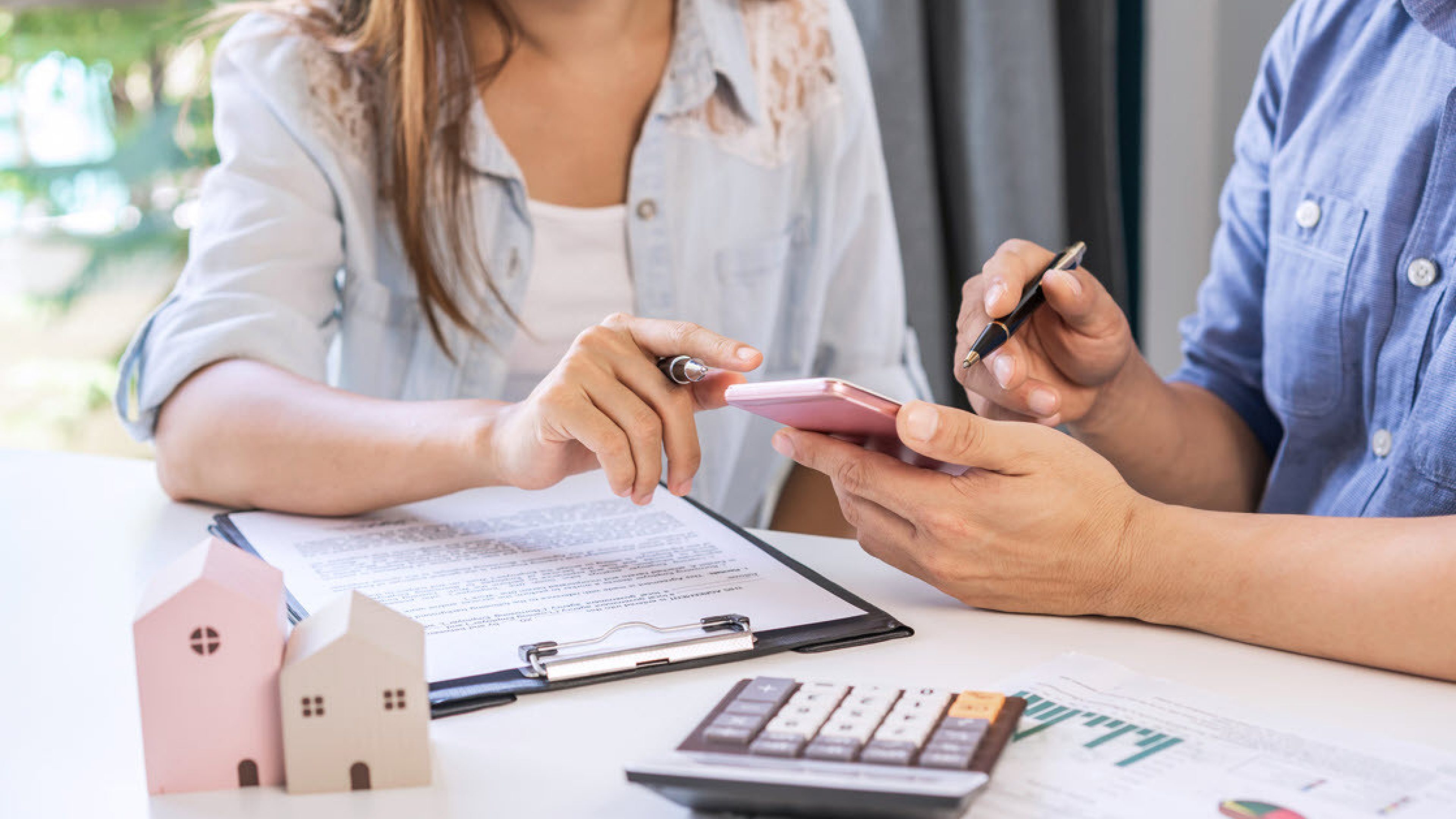 Une femme et un homme sont assis à une table et calculent le financement du logement en propriété. Sur la table se trouvent des documents, une calculatrice et deux maisons modèles.