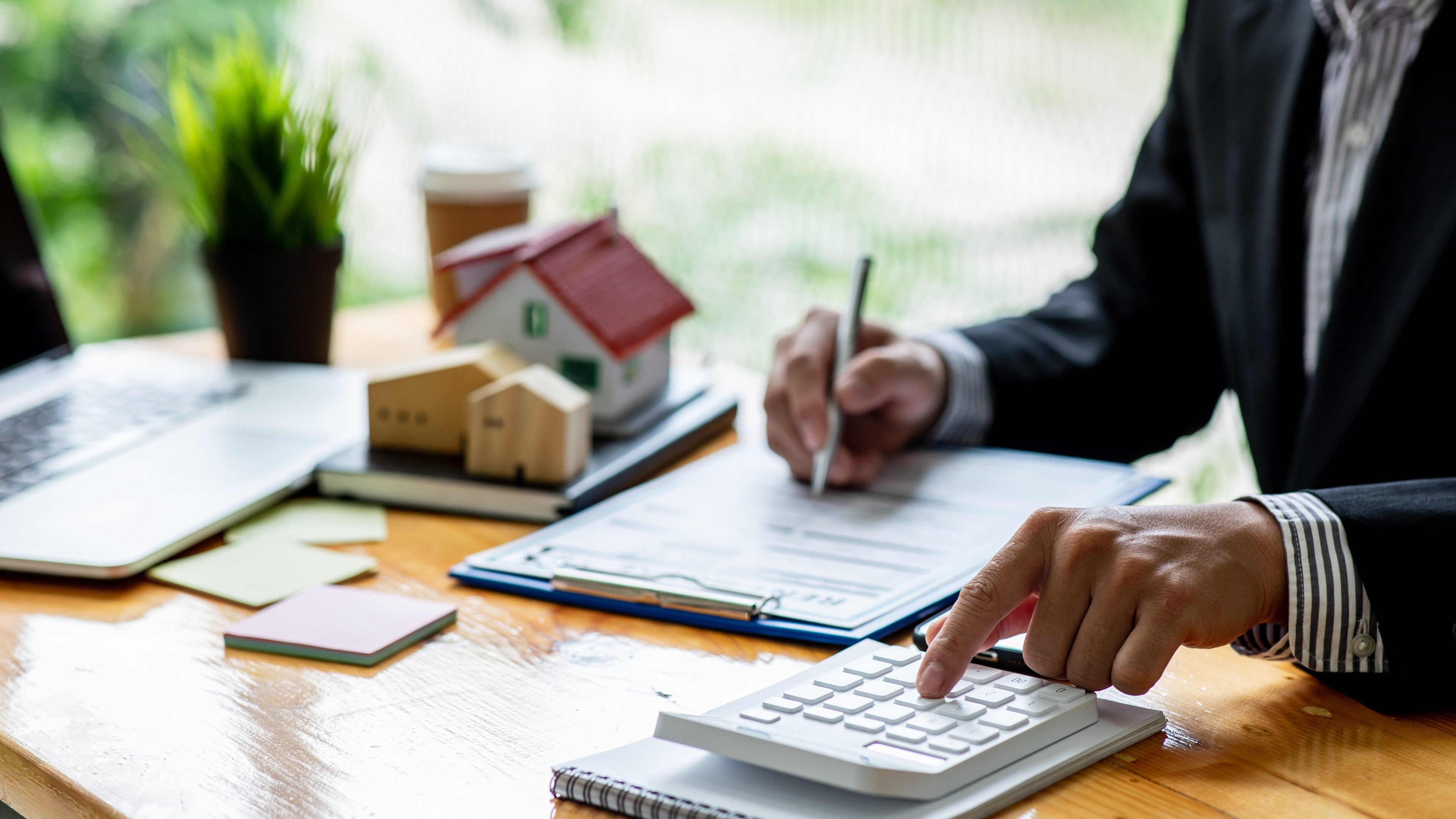 Un conseiller en immobilier est assis à une table et calcule quelque chose sur sa calculatrice. Sur la table se trouvent des documents et une maison modèle jaune.