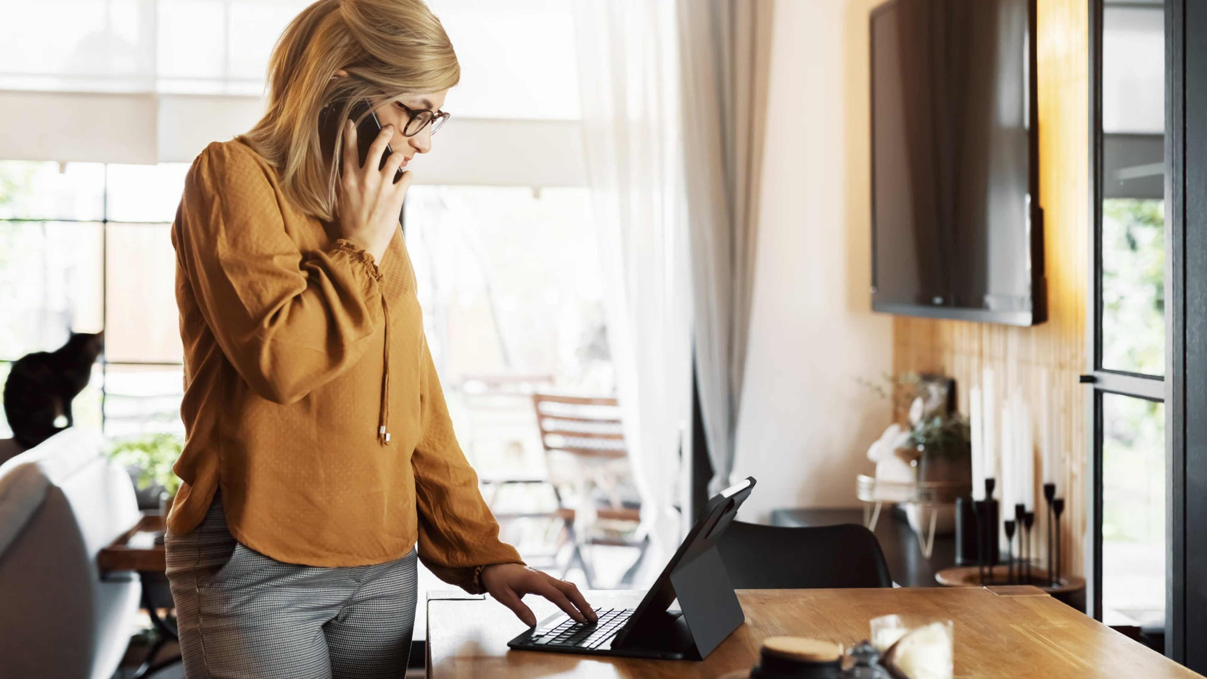 Une femme souriante se tient devant une tablette dans le salon, téléphone et effectue son travail en home office. En arrière-plan, on voit une télévision et un chat noir.